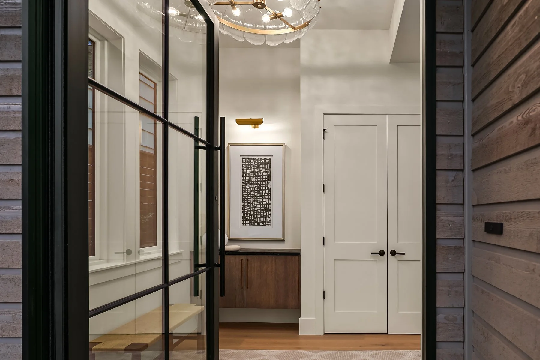 View of a modern entryway with white walls, a wooden cabinet, abstract wall art, and a chandelier, seen through a black-framed glass door.