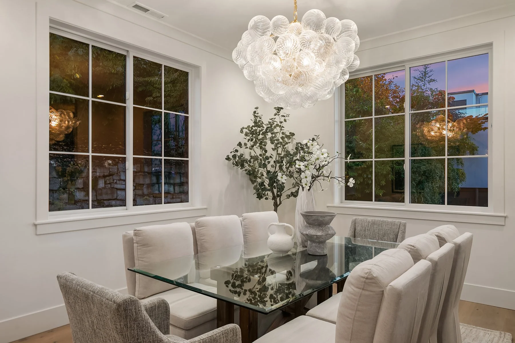 A dining room with a glass table, white upholstered chairs, a large ceiling chandelier made of glass bubbles, a large potted plant, two vases on the table, and large windows showing a twilight sky outside.