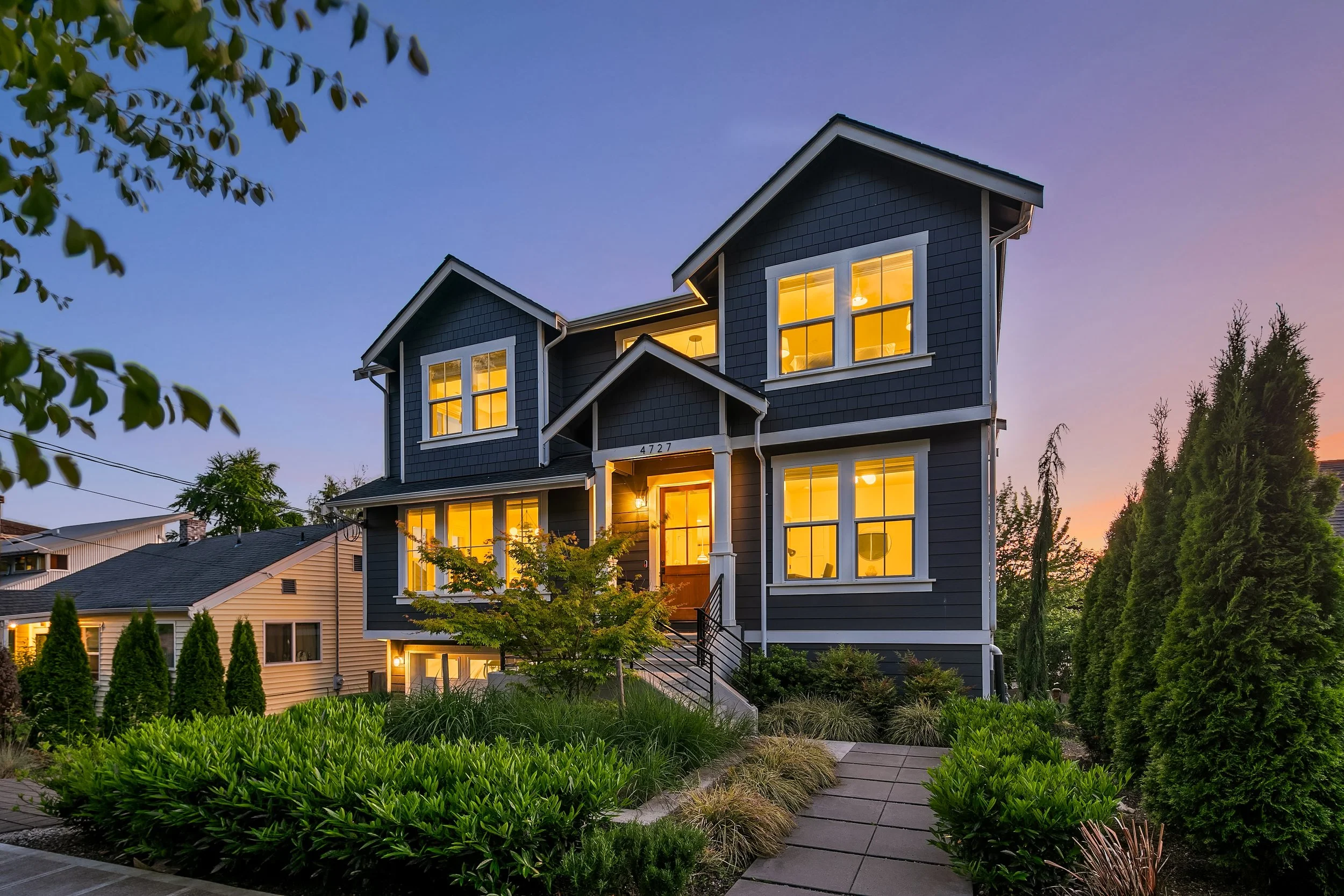 A dark blue, three-story house with white trim, lit from inside during sunset, with a front yard filled with green bushes, trees, and a paved walkway leading to the front steps.