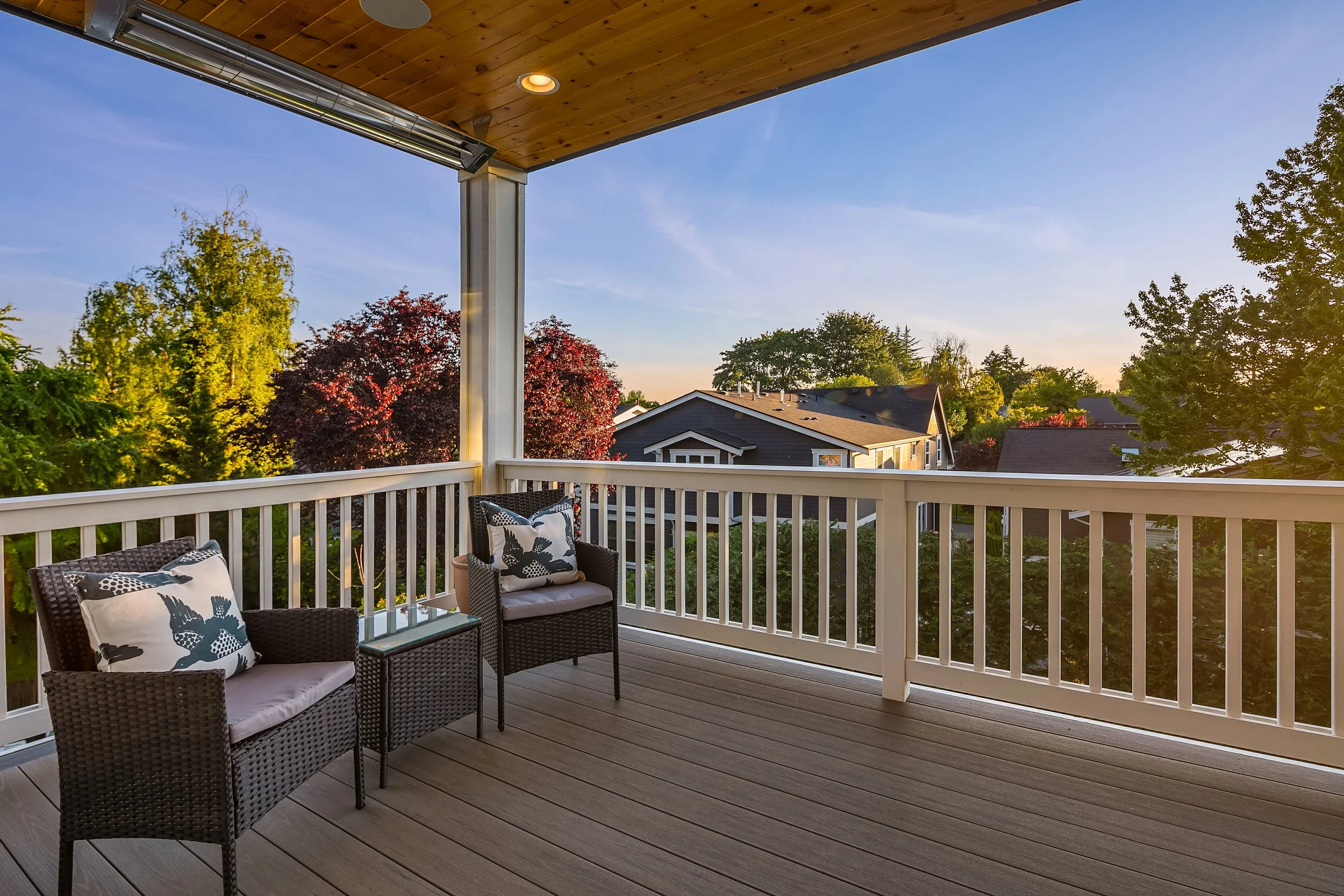 Empty balcony with two wicker chairs with cushions, a small glass table, white railing, wooden overhang, and a view of trees and houses at sunset.