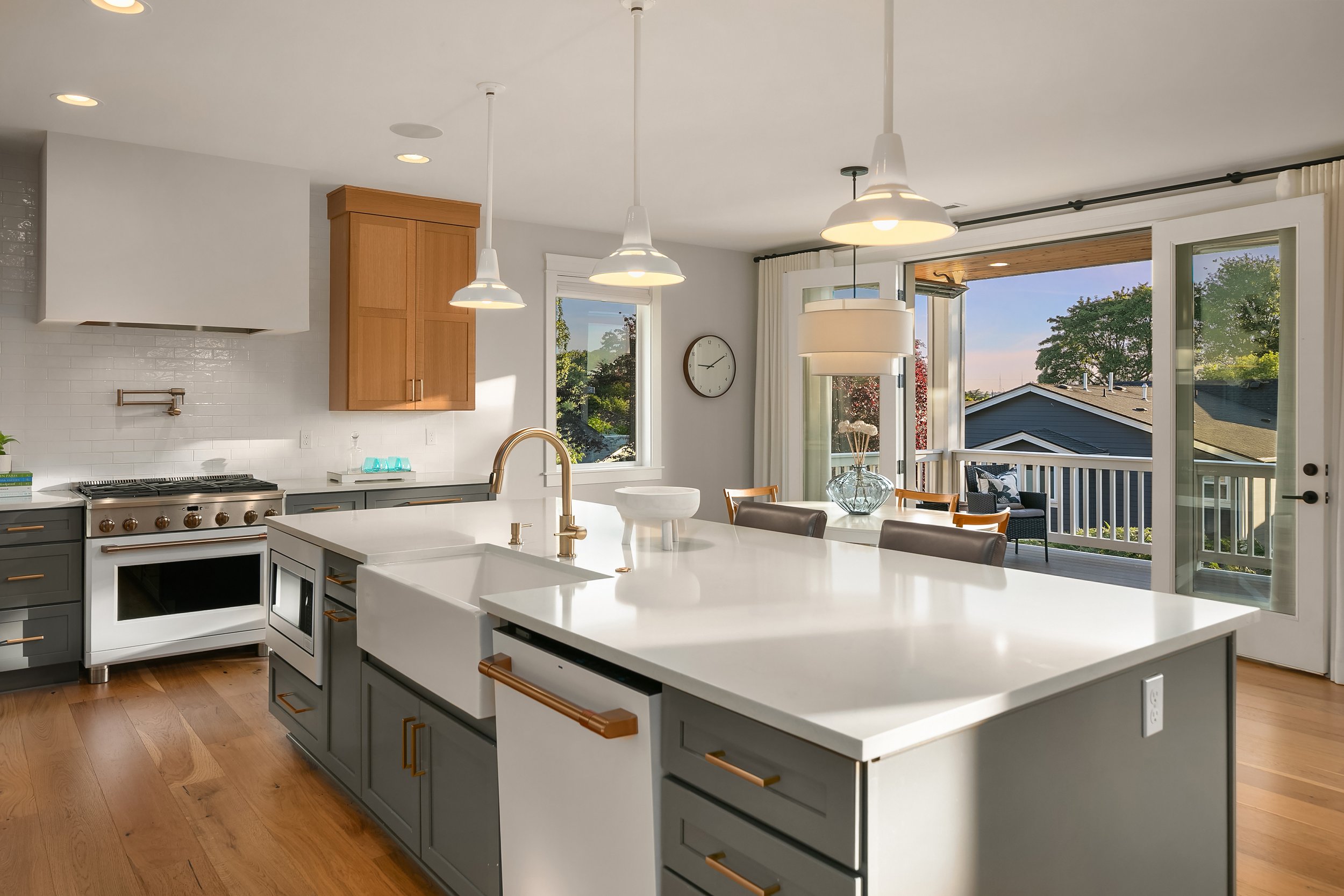 Modern kitchen with white island, gray cabinets, wooden bar handles, stainless steel appliances, and large windows overlooking a backyard with trees and a blue sky.