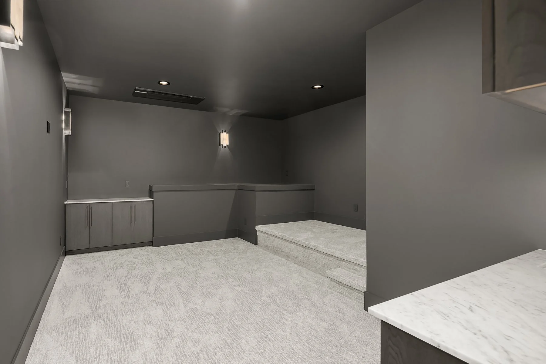 Empty room with gray walls, carpeted floor, and recessed lighting, possibly a basement or lounge area.