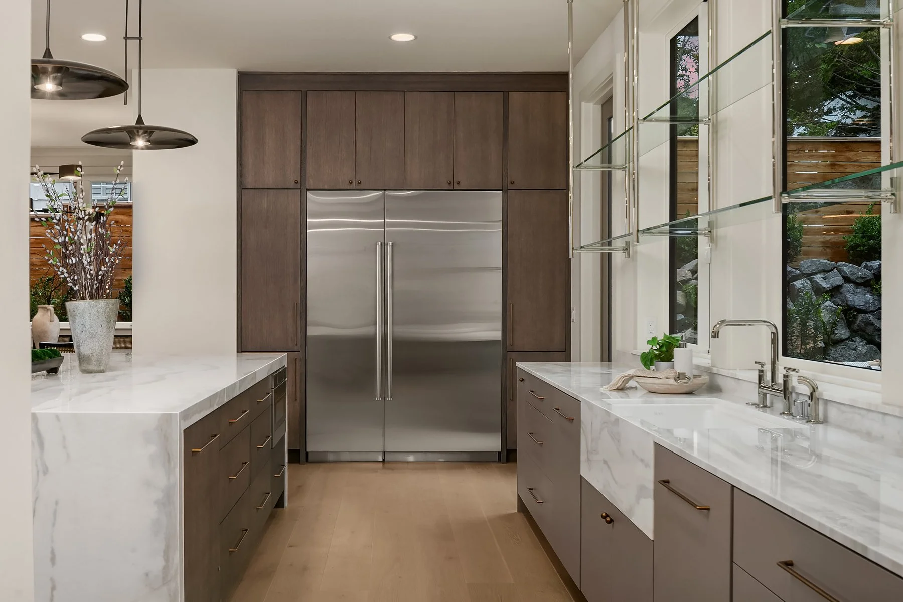 Modern kitchen with marble countertops, brown cabinetry, and large stainless steel refrigerator. Two black pendant lights hanging above the counter, with a window view of greenery outside.