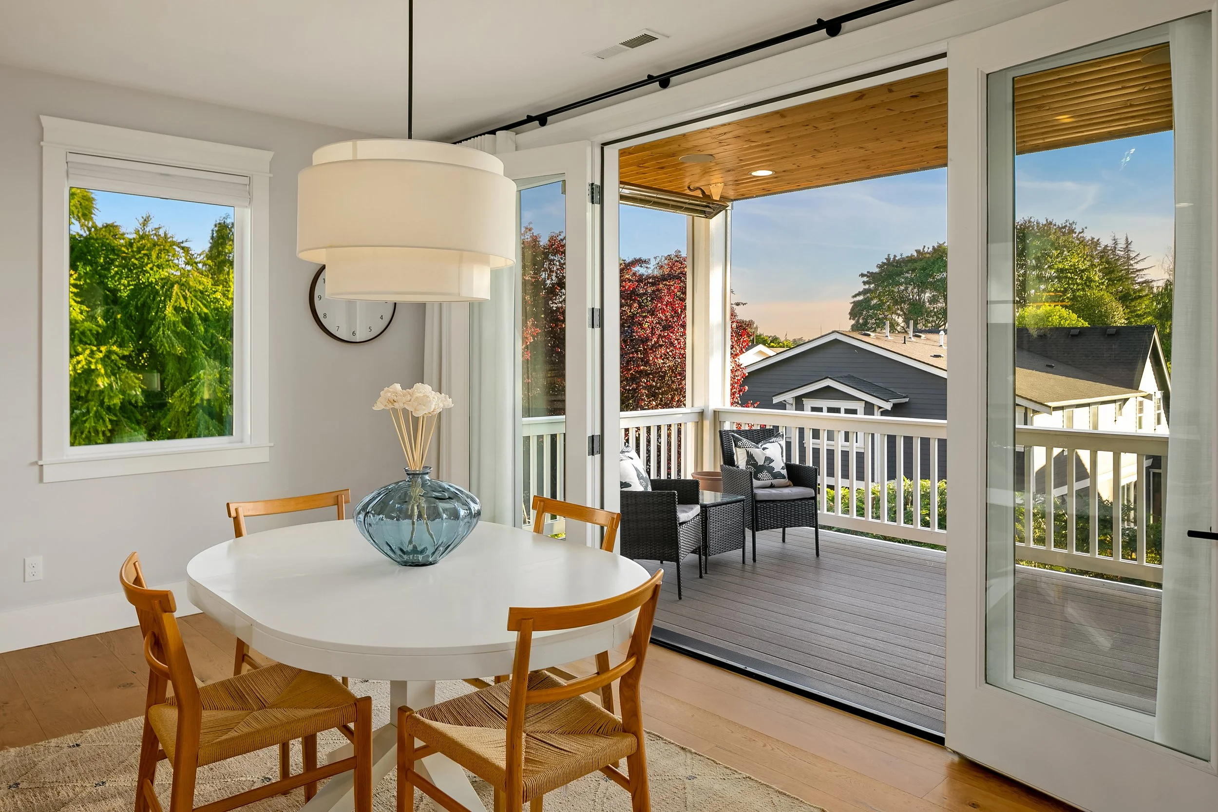 Dining room with a round white table, four wooden chairs, a large pendant light, a glass vase with white flowers, a window, and an open door leading to a balcony with two black chairs and scenic view of trees and neighboring houses.