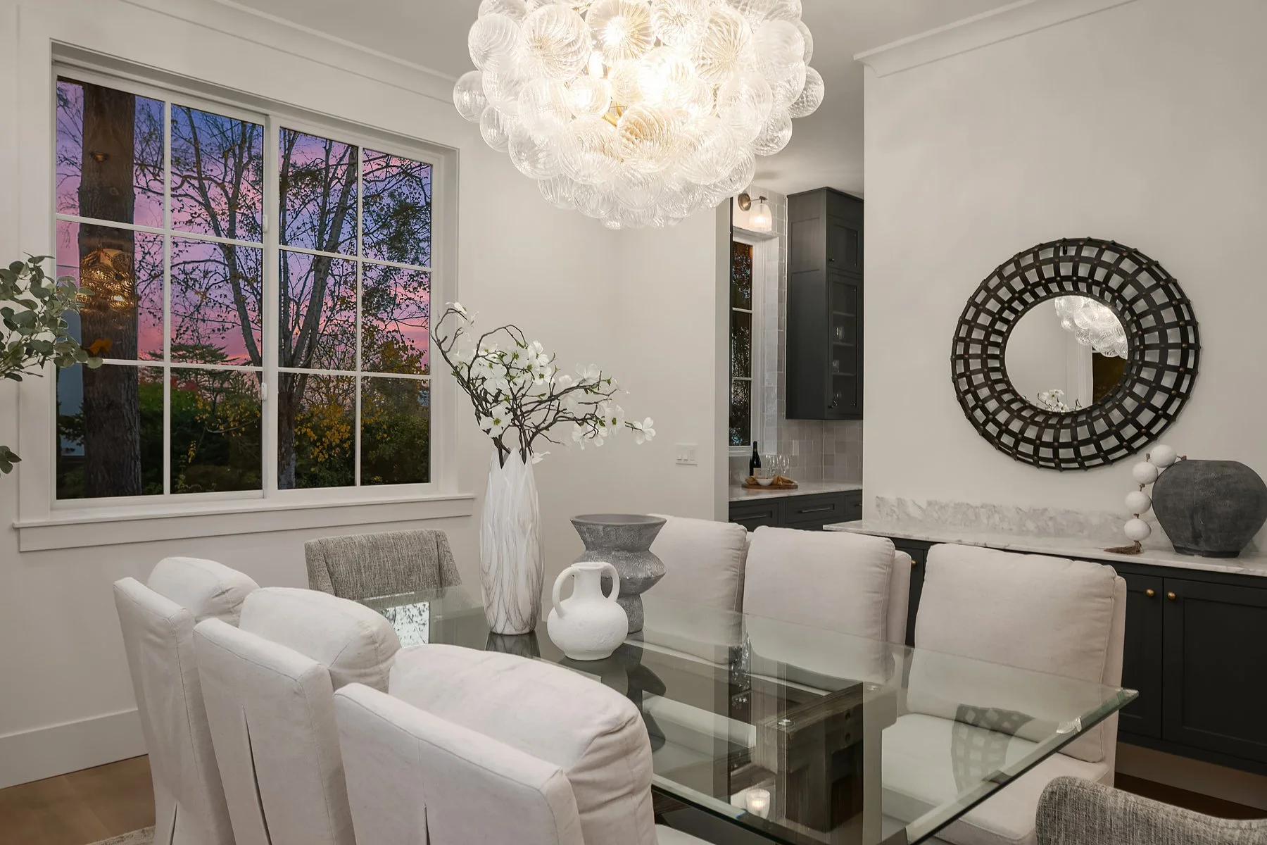 A modern dining room featuring a glass-top table surrounded by white and gray upholstered chairs. A large, spherical chandelier hangs above. The room has a window showing a sunset sky with trees outside, and decor includes a tall white vase with bran