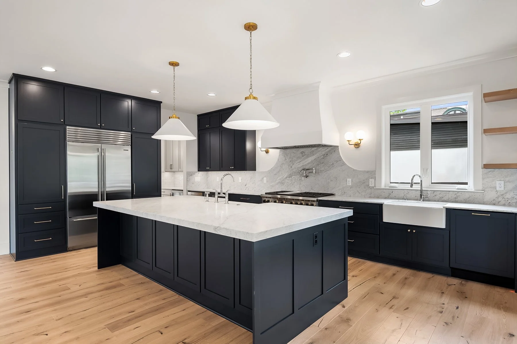 Modern kitchen with navy blue cabinets, white marble countertops, a large island, and hardwood flooring. There are two pendant lights above the island, a stainless steel refrigerator, stovetop, and double sink under a window.