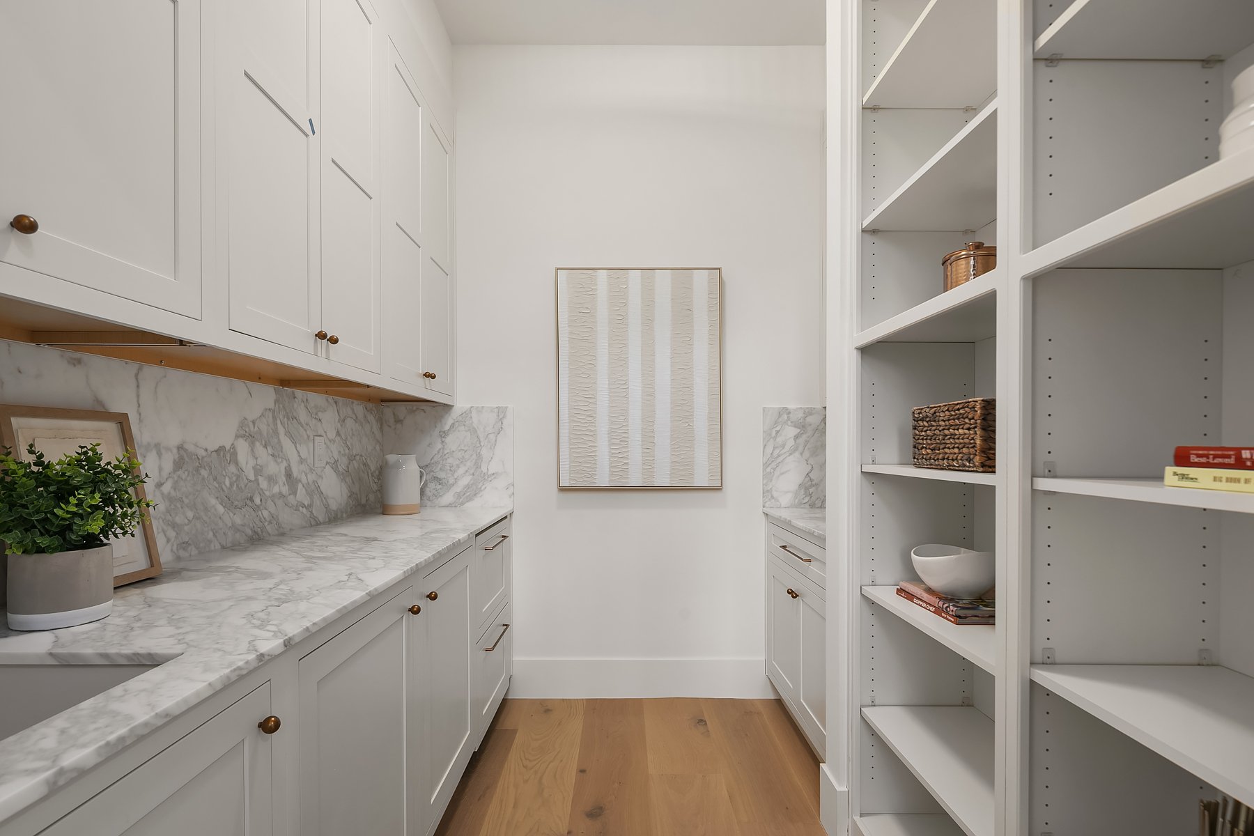 A clean, organized pantry with white cabinetry, marble countertops, and wooden floors. The shelves hold baskets, books, and small bowls, with a decorative artwork on the back wall.