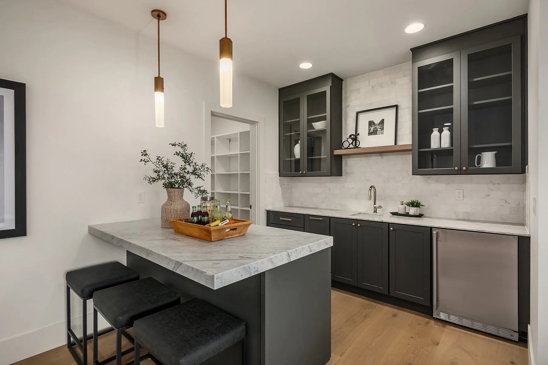 Modern kitchen with dark gray cabinetry, white marble countertops, and a small bar area with three black stools. There are two pendant lights hanging from the ceiling and a small potted plant on the countertop.