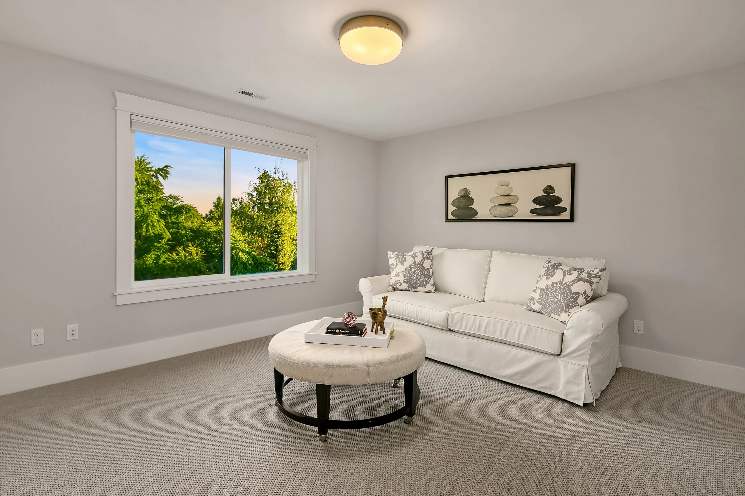 Living room with a white sofa, decorative pillows, framed art of stacked stones, round ottoman with tray of decor, window overlooking green trees, beige carpet, ceiling light
