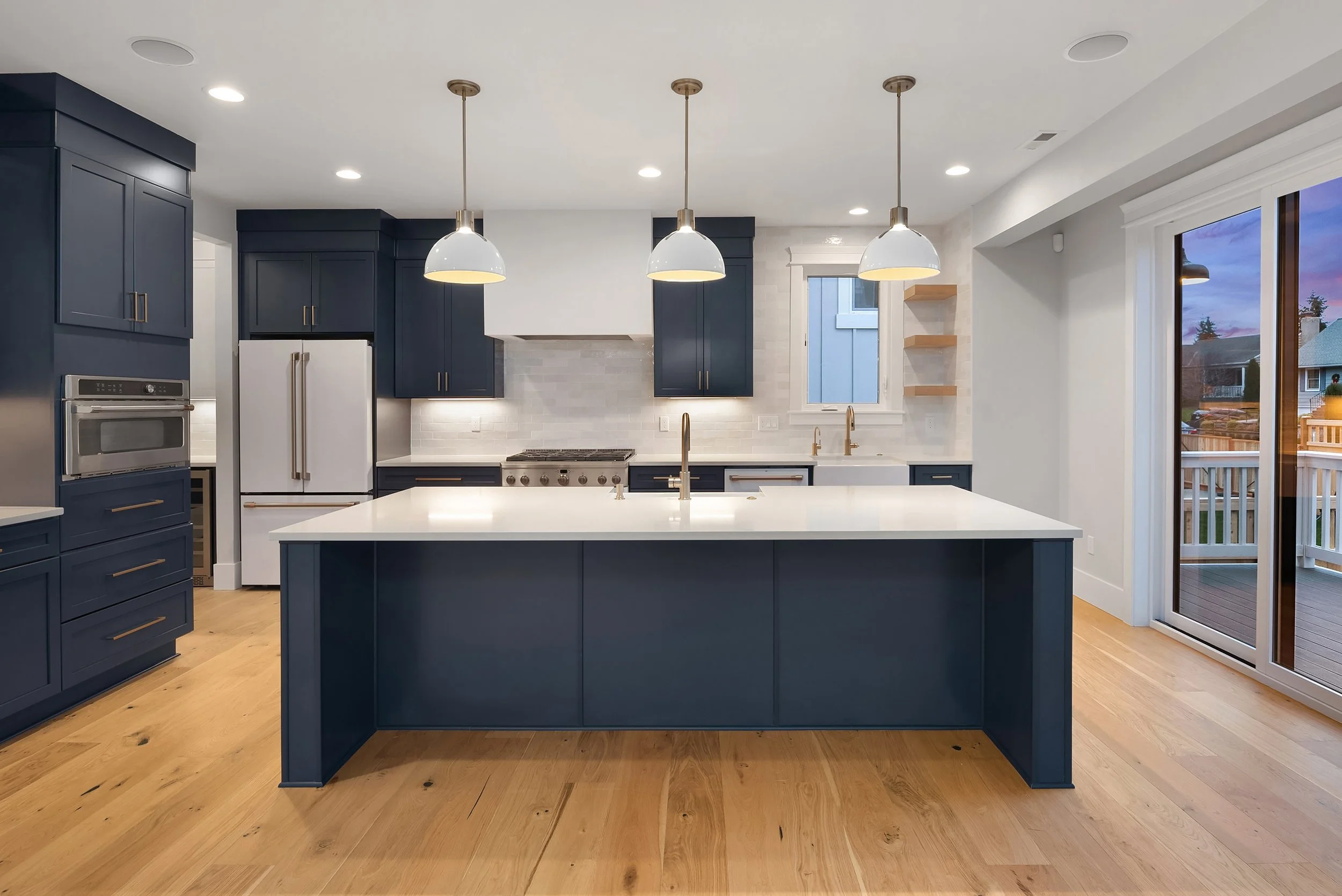 Modern kitchen with blue cabinets, white countertop island, stainless steel appliances, light wood flooring, and large sliding glass door leading to an outdoor deck.