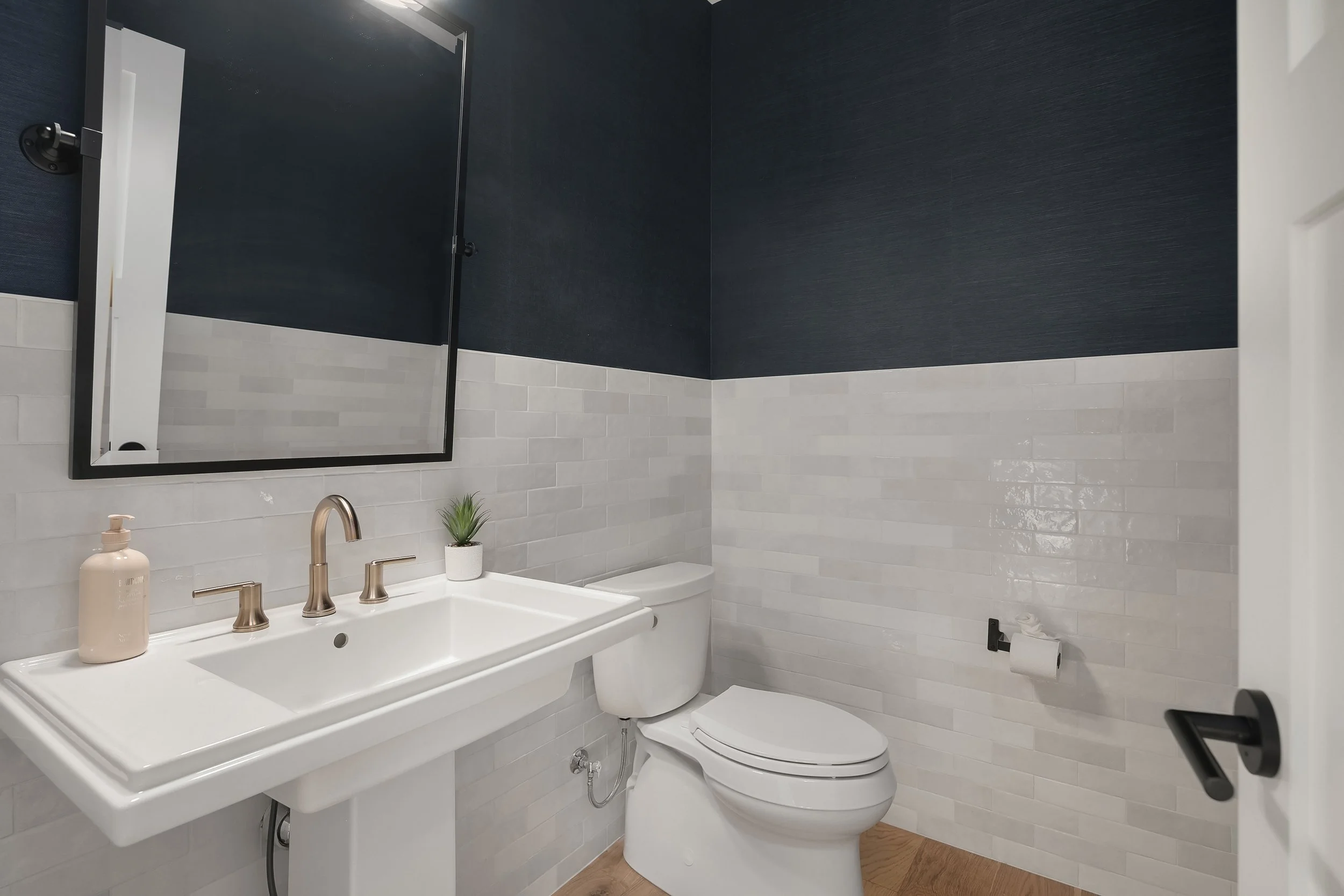 A modern bathroom with a white pedestal sink, a toilet, and a large mirror above the sink. The walls are painted dark blue on the upper half with white subway tiles on the lower half. There is a small green plant in a white pot on the sink and a soap