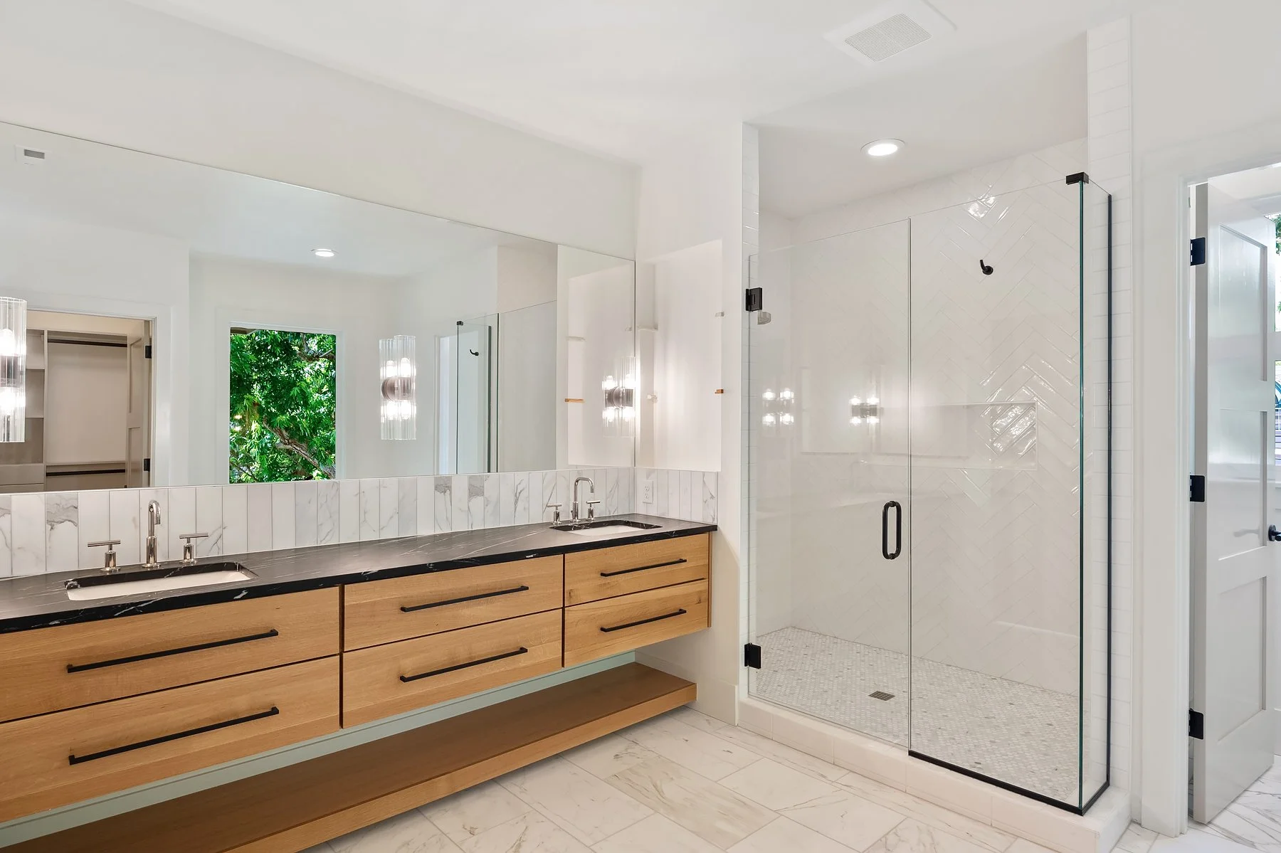 Modern bathroom with double vanity, black countertops, wooden drawers, large mirror, walk-in glass shower with white herringbone tiles, and window showing greenery outside.