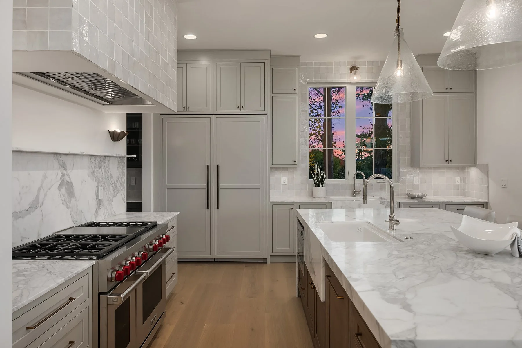 Modern kitchen with white marble countertops, gray cabinetry, and a large window showing a sunset with pink and purple sky and trees outside.