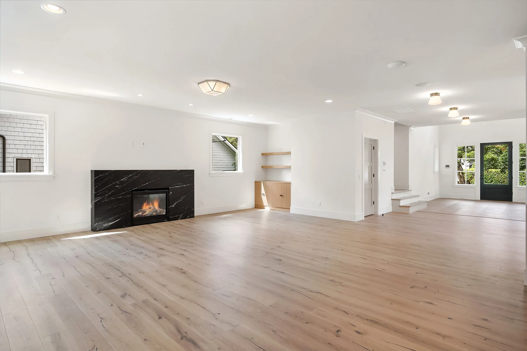 Empty living room with white walls, light wooden flooring, a black marble fireplace, and multiple windows allowing natural light.