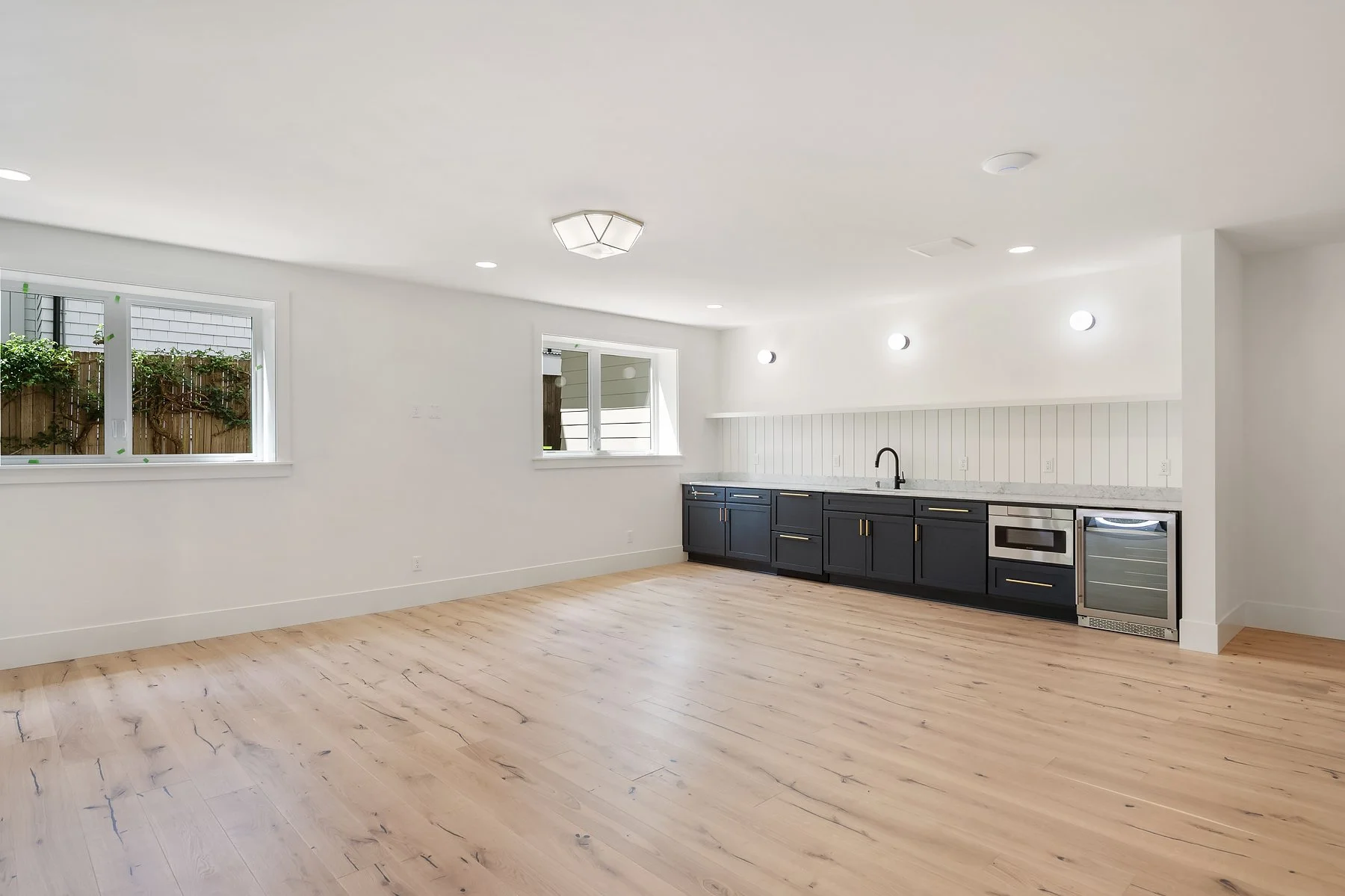 Empty kitchen with white walls, light wooden floors, black lower cabinets, a marble countertop, a small oven, a sink, and three windows overlooking an outdoor space.