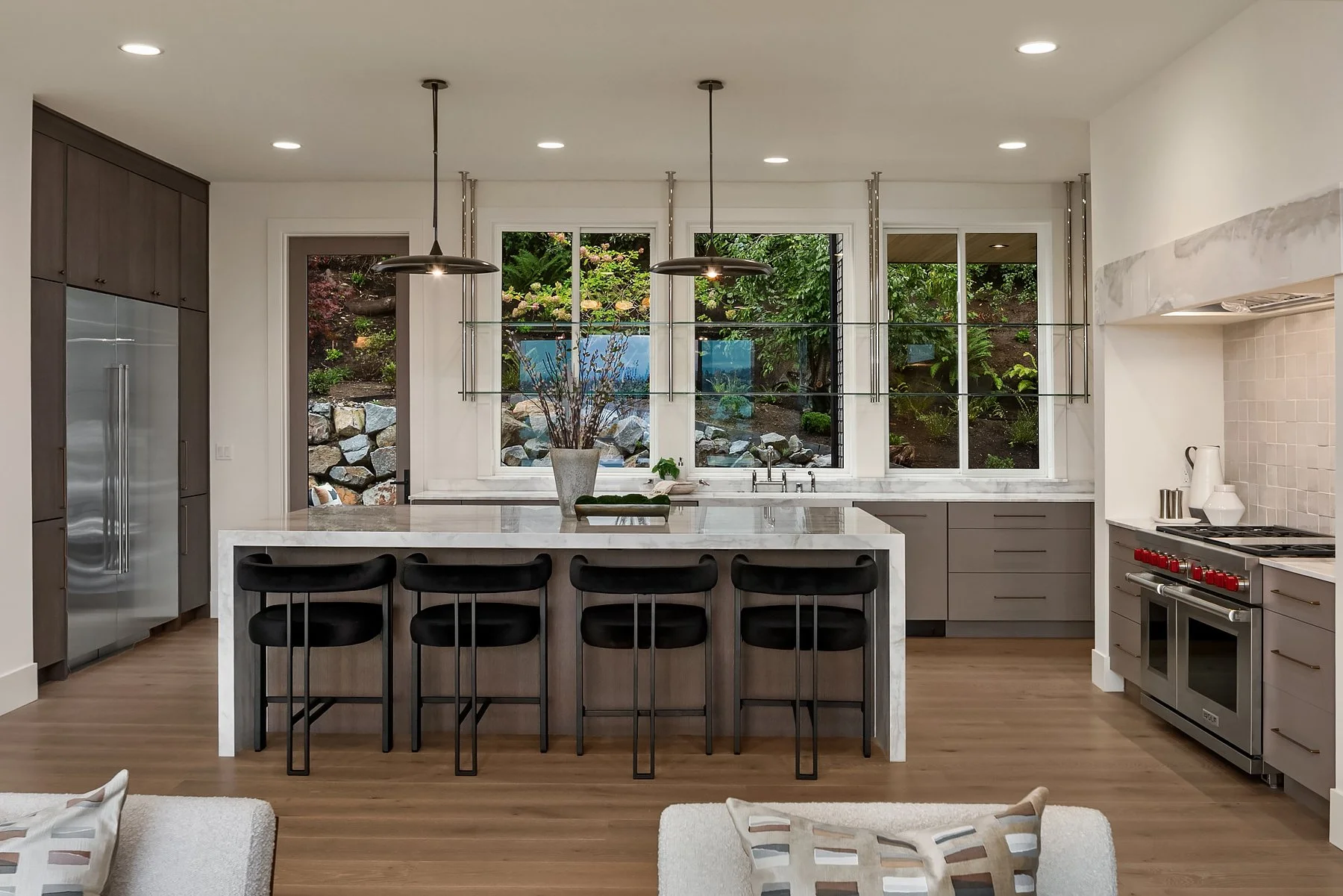 Modern kitchen with gray cabinets, a white marble island, black bar stools, large windows, and a view of a landscaped backyard.