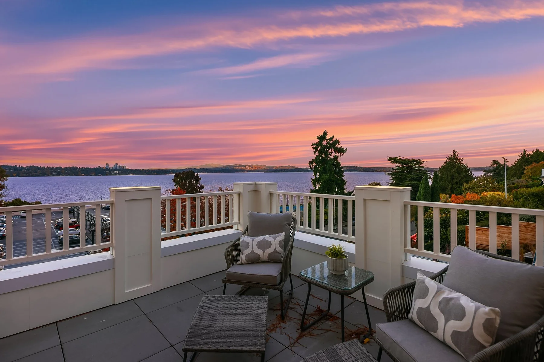 View of a lake at sunset from a balcony with outdoor furniture and trees.
