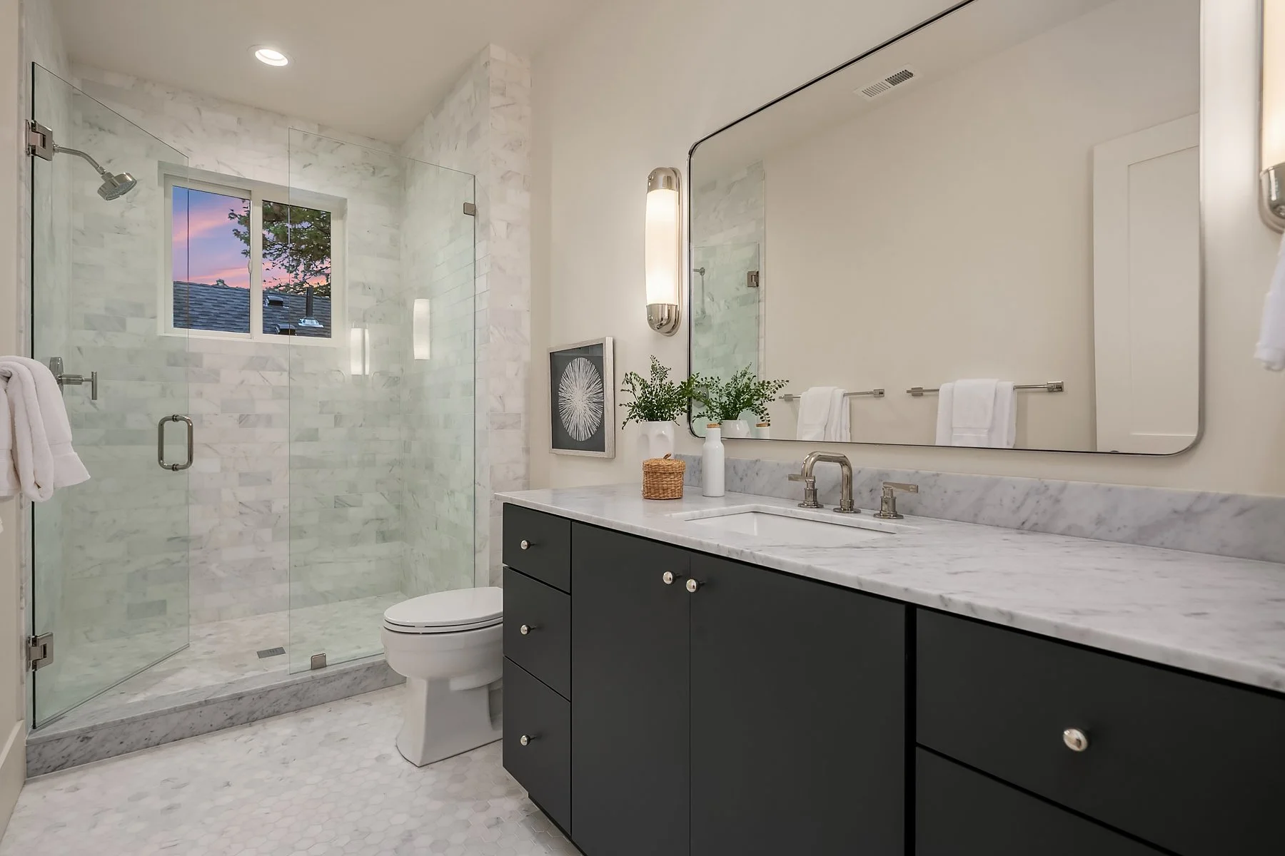 Modern bathroom with a marble shower, a dark vanity with a marble countertop, a large mirror, potted plants, and a window showing a sunset