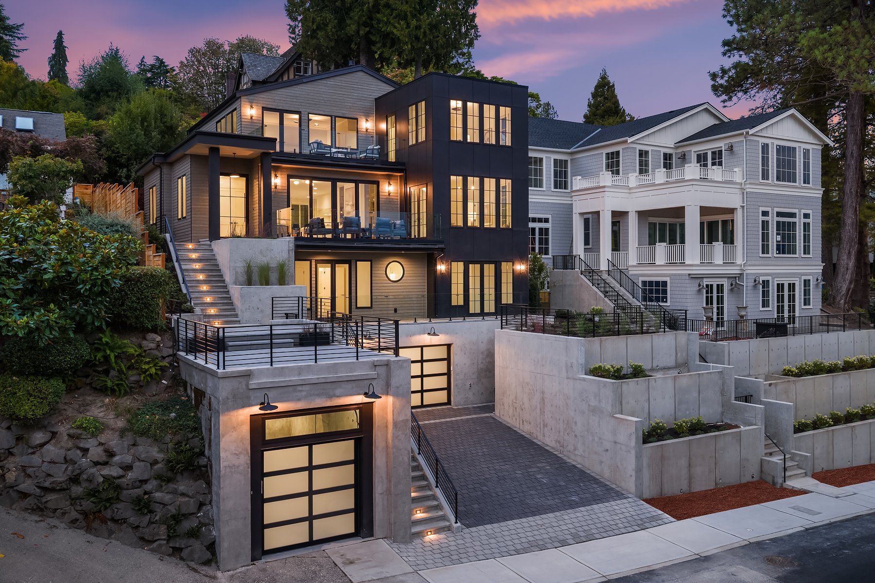 Modern multi-story house on a hillside at dusk with illuminated interior lights, featuring multiple balconies, large windows, concrete stairs, and landscaped tiered garden with plants and hardscaping.