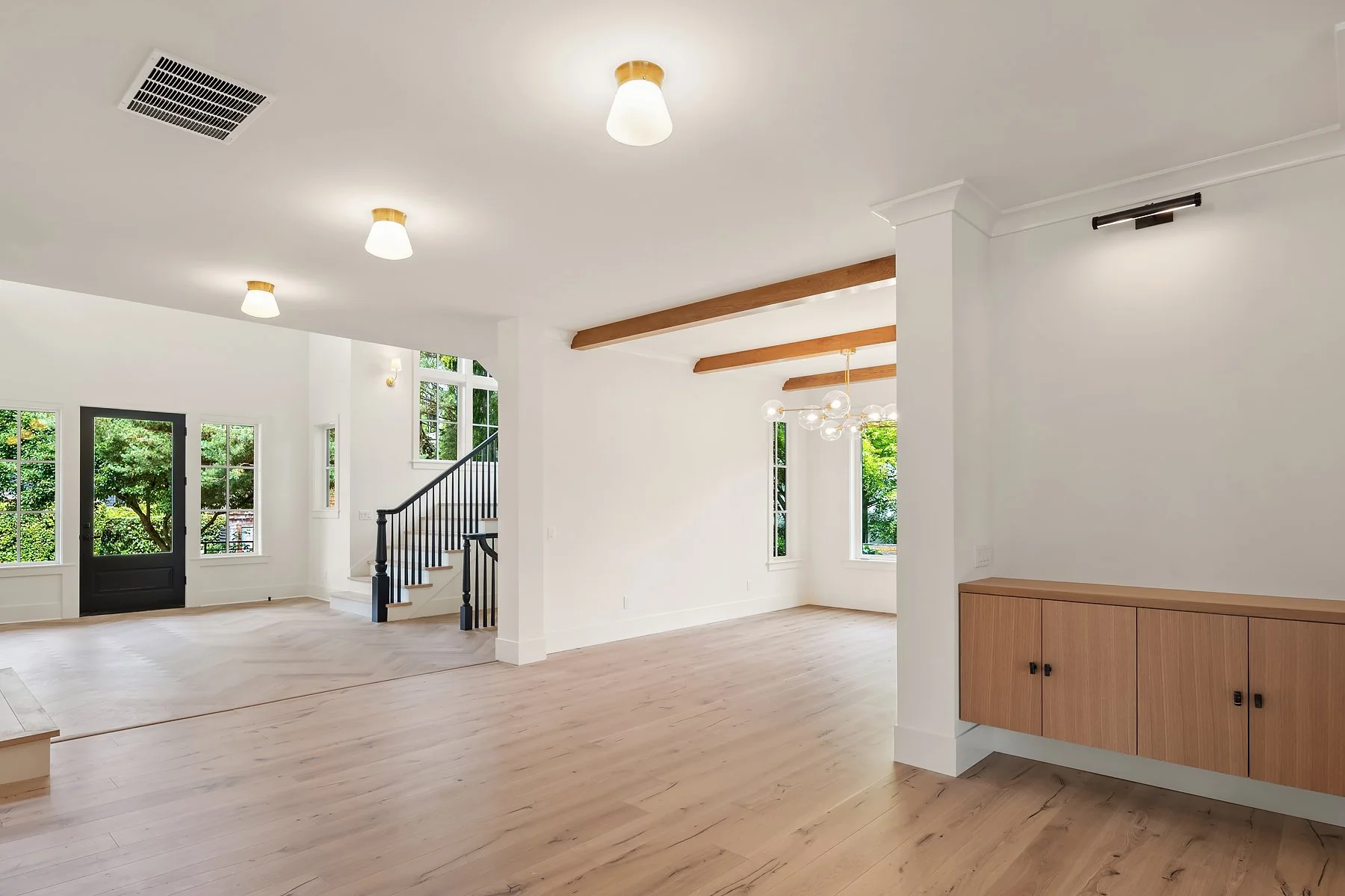 Open living area with wood flooring, large windows, a black front door, staircase with black railing, and modern light fixtures on white walls.