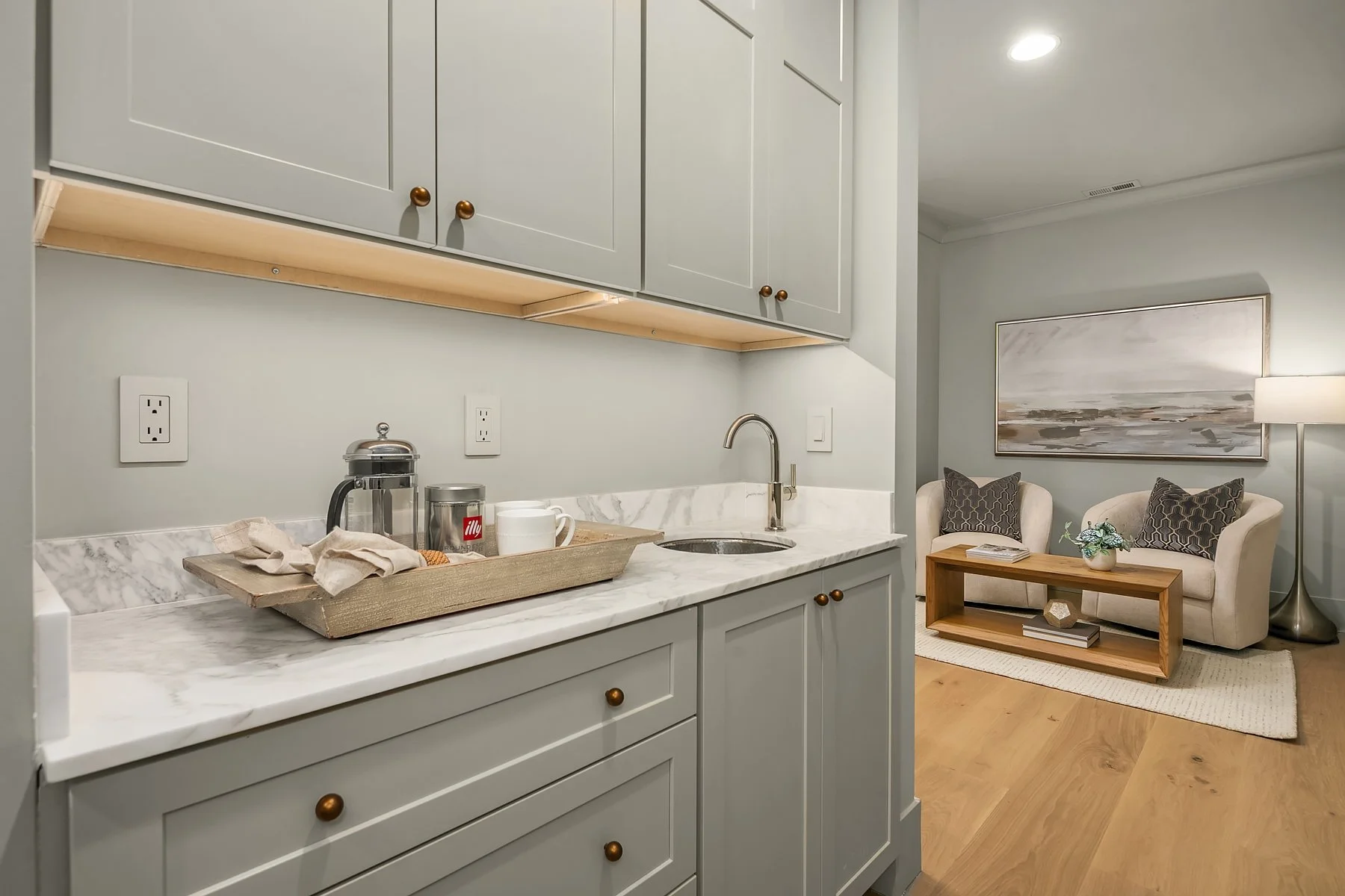 View of a small kitchen area with light gray cabinetry, marble countertops, and a single basin under-mount sink. There are two electrical outlets on the wall above the counter. In the background, a cozy seating area with a white sofa, two patterned p