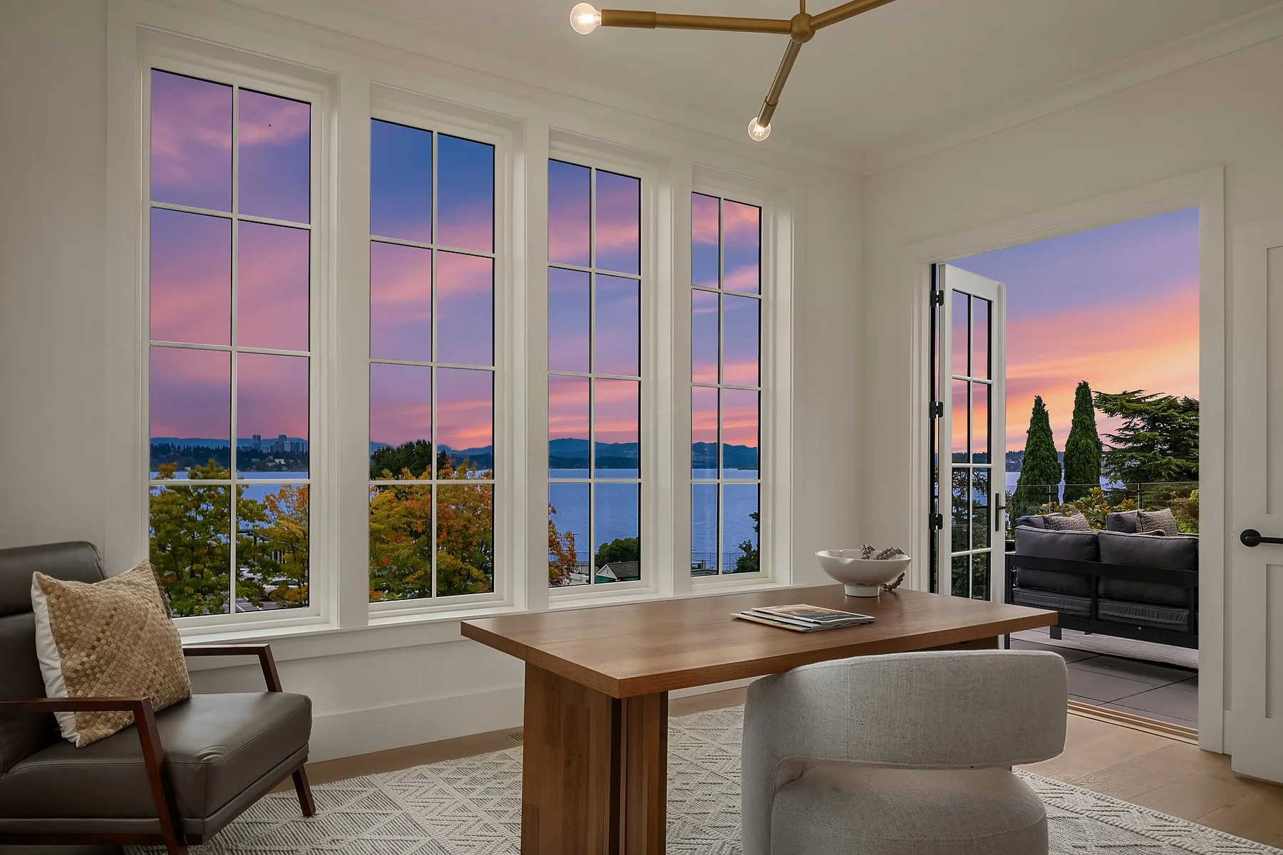 Living room with large windows and a door opening to a balcony, overlooking a body of water and a sunset sky, with trees and distant cityscape, decorated with a wooden table, chairs, and a ceiling light.