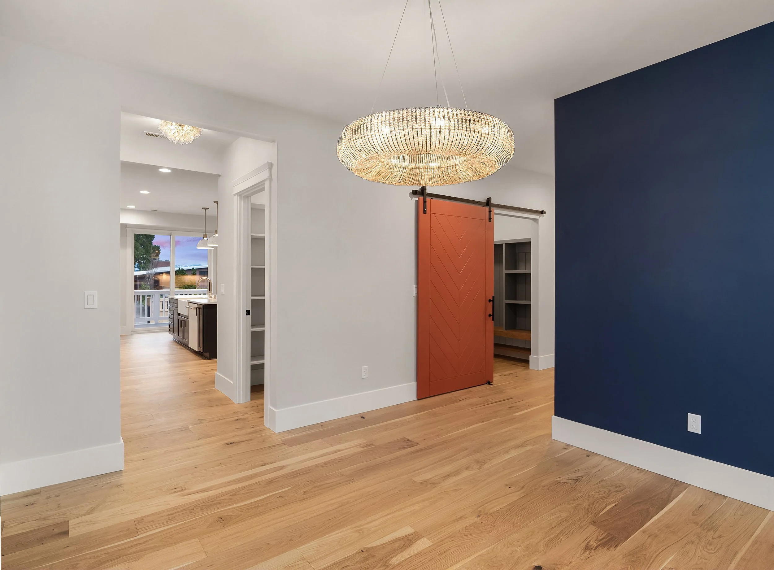 Modern home interior with light wood floors, a blue accent wall, and a round chandelier. A sliding barn door with a chevron pattern in rust color is partially open, revealing shelving inside. The space opens into a kitchen area with large windows and