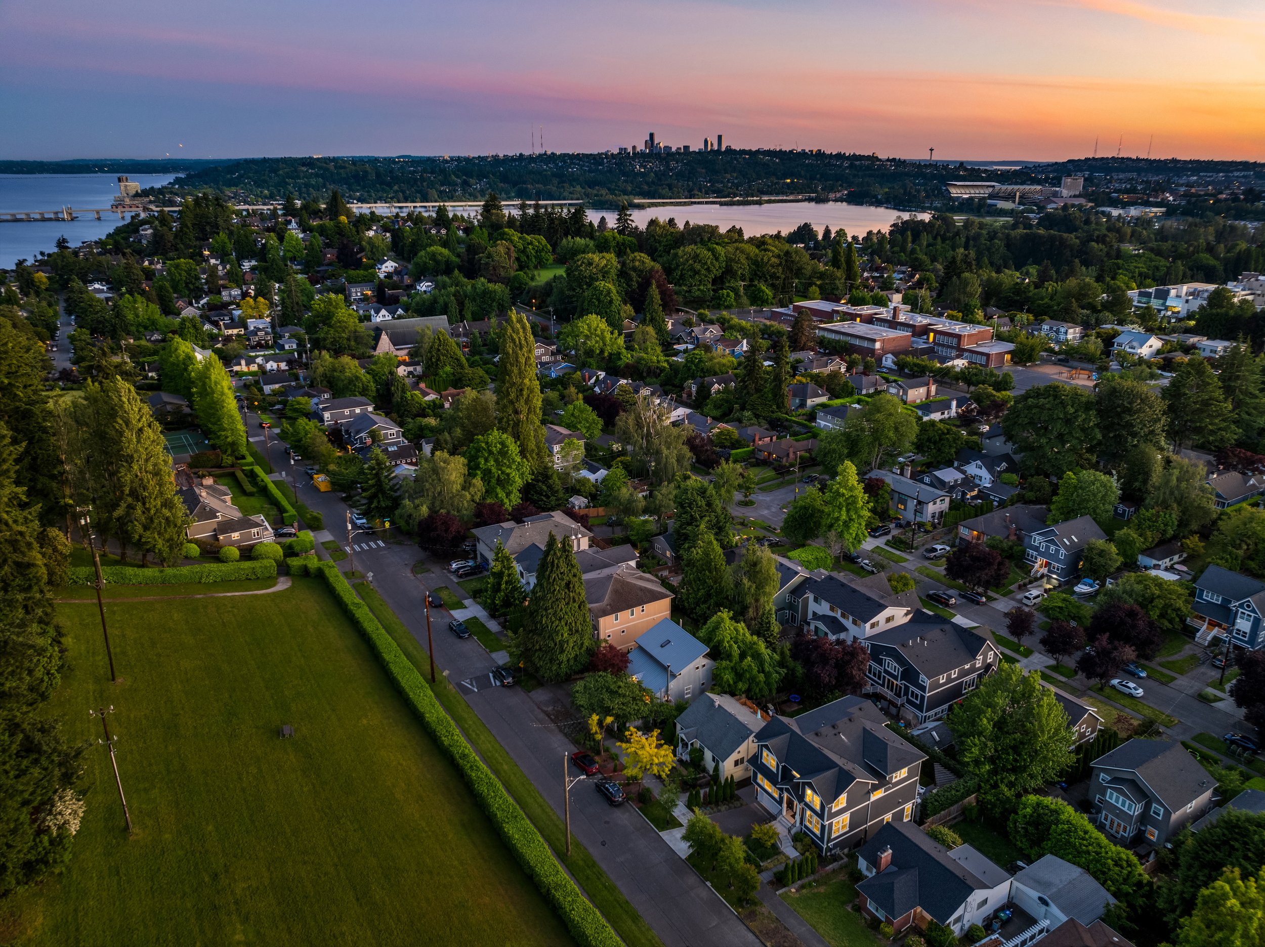 Aerial view of a suburban neighborhood at sunset with houses, trees, and a river in the background.