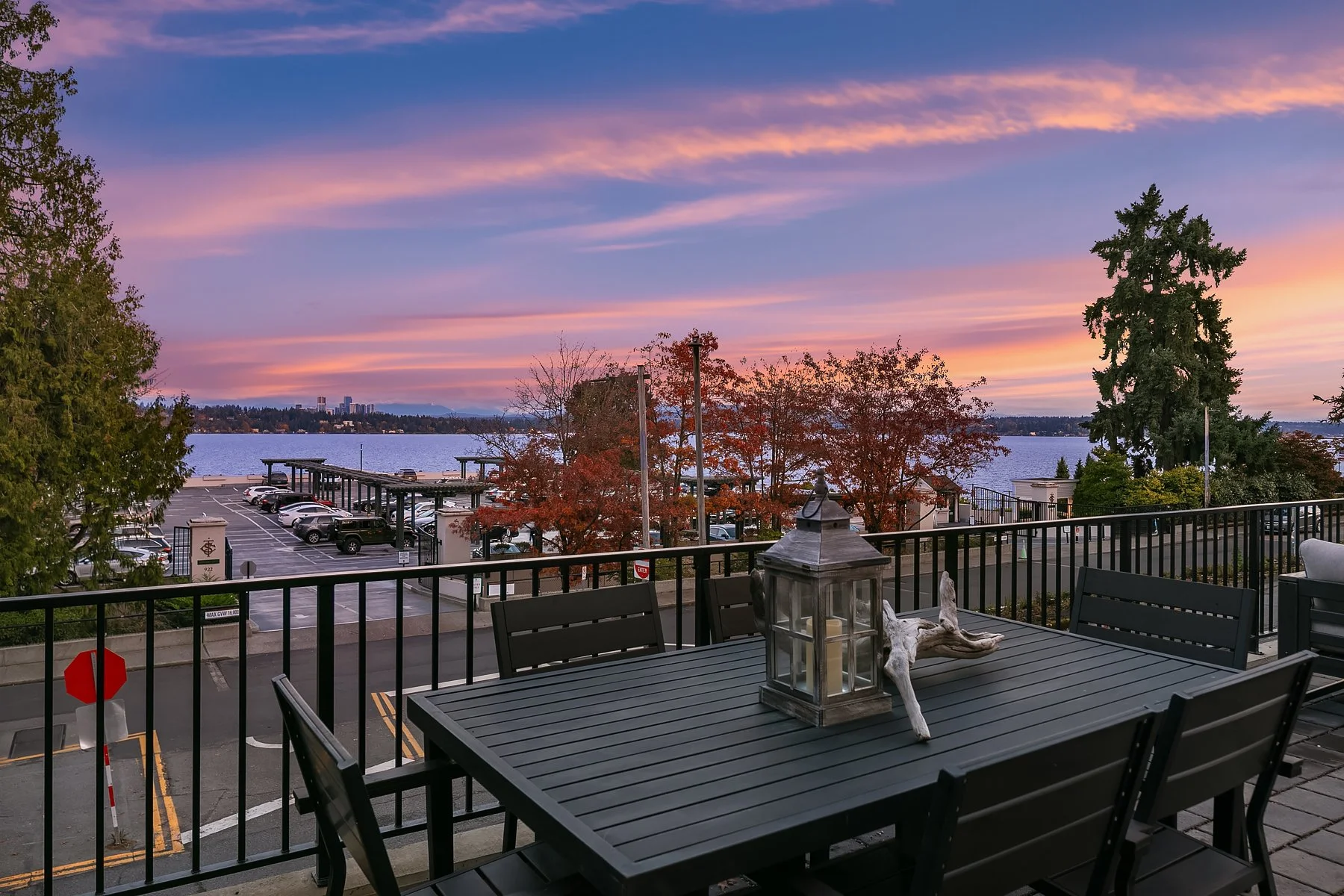 A balcony overlooking a parking lot and a body of water during a colorful sunset sky. There is a black patio table and chairs with a lantern and decorative driftwood on the table.