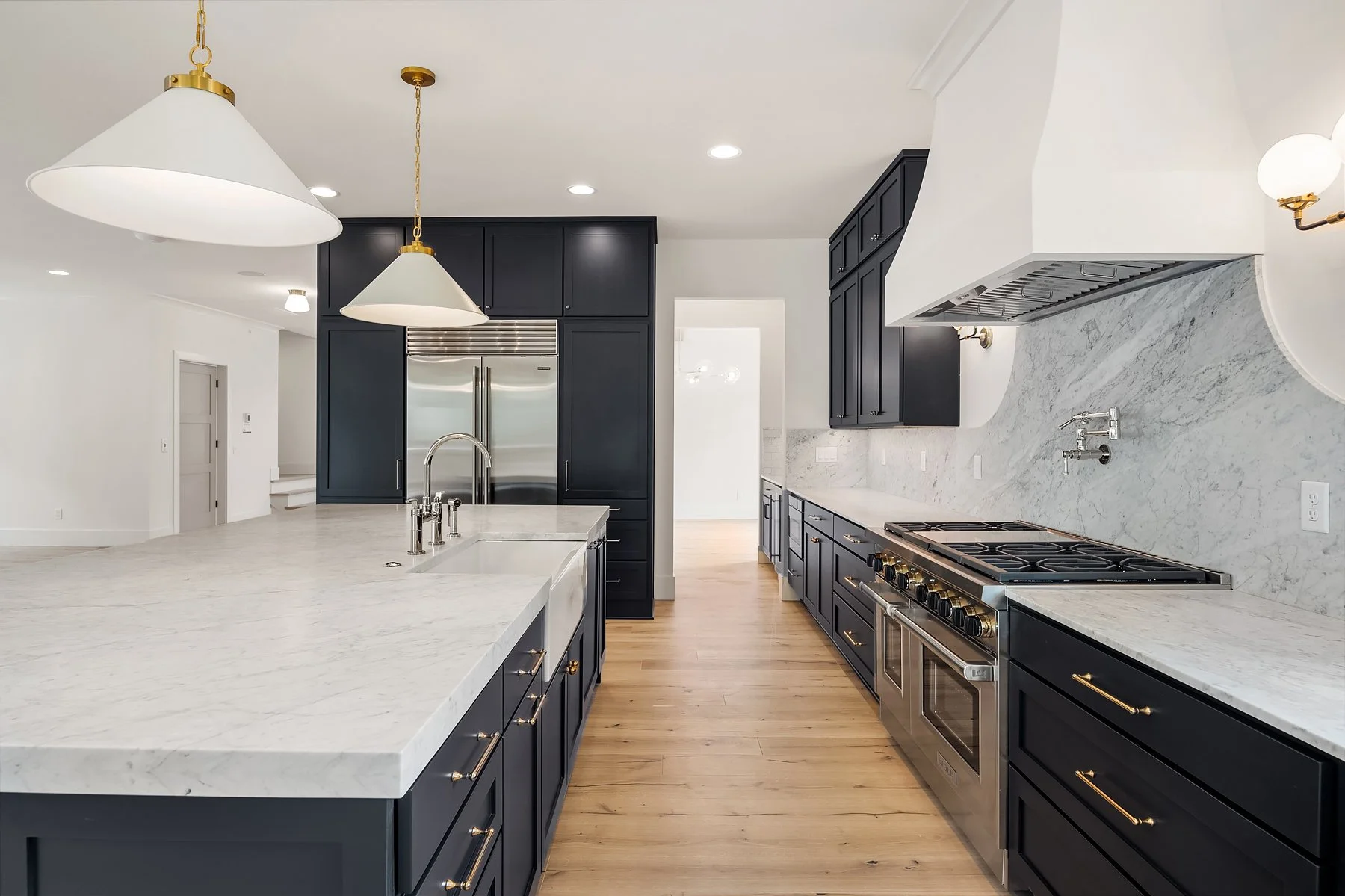 Modern kitchen with white marble countertops, black cabinets with gold handles, stainless steel appliances, and a wooden floor, illuminated by pendant and ceiling lights.