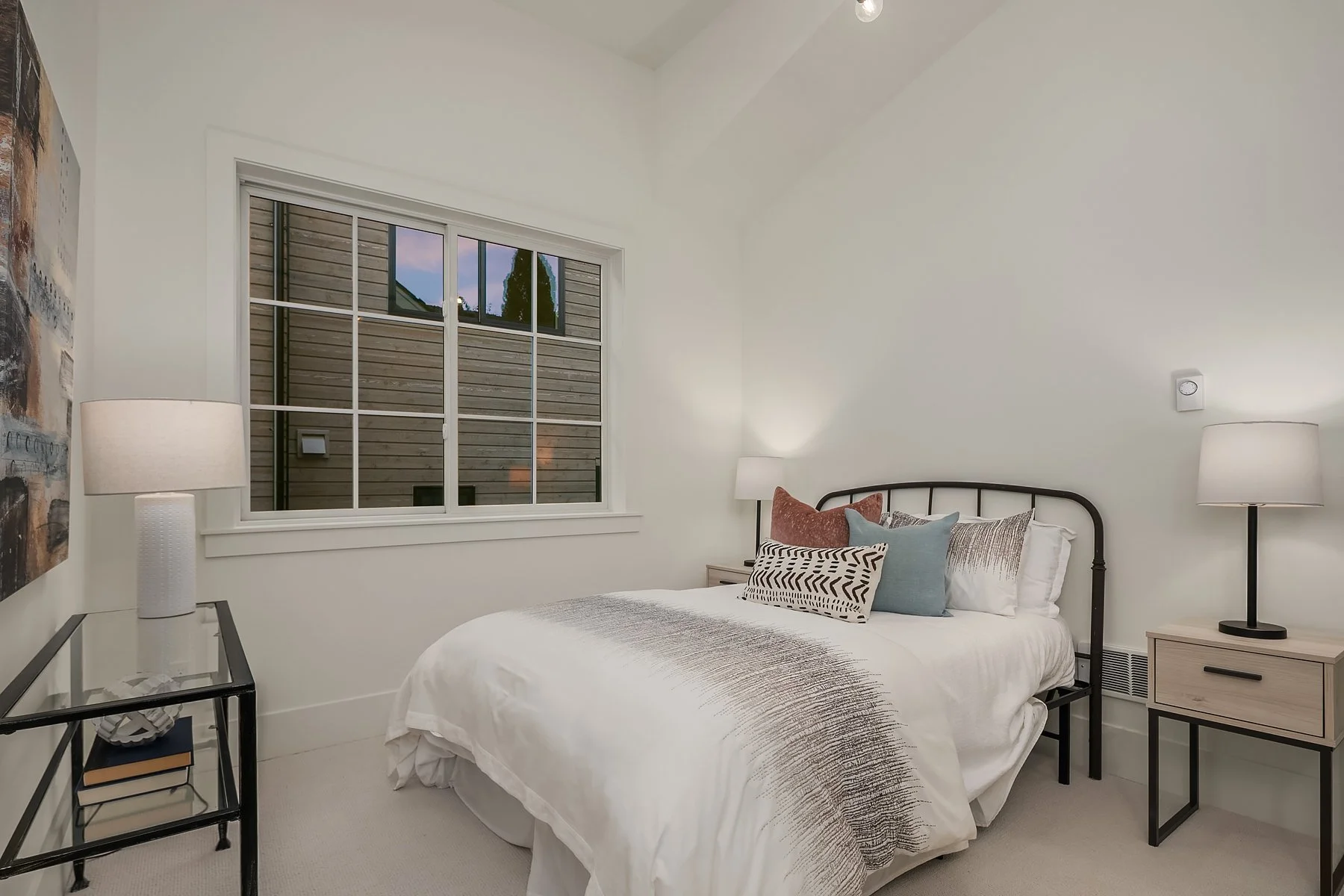 Cozy bedroom with white walls, a bed with white and patterned bedding, and multiple pillows including rust, blue, and black-and-white designs. Two bedside tables with black frame and light wood drawers hold white lamps. A window shows exterior wood s