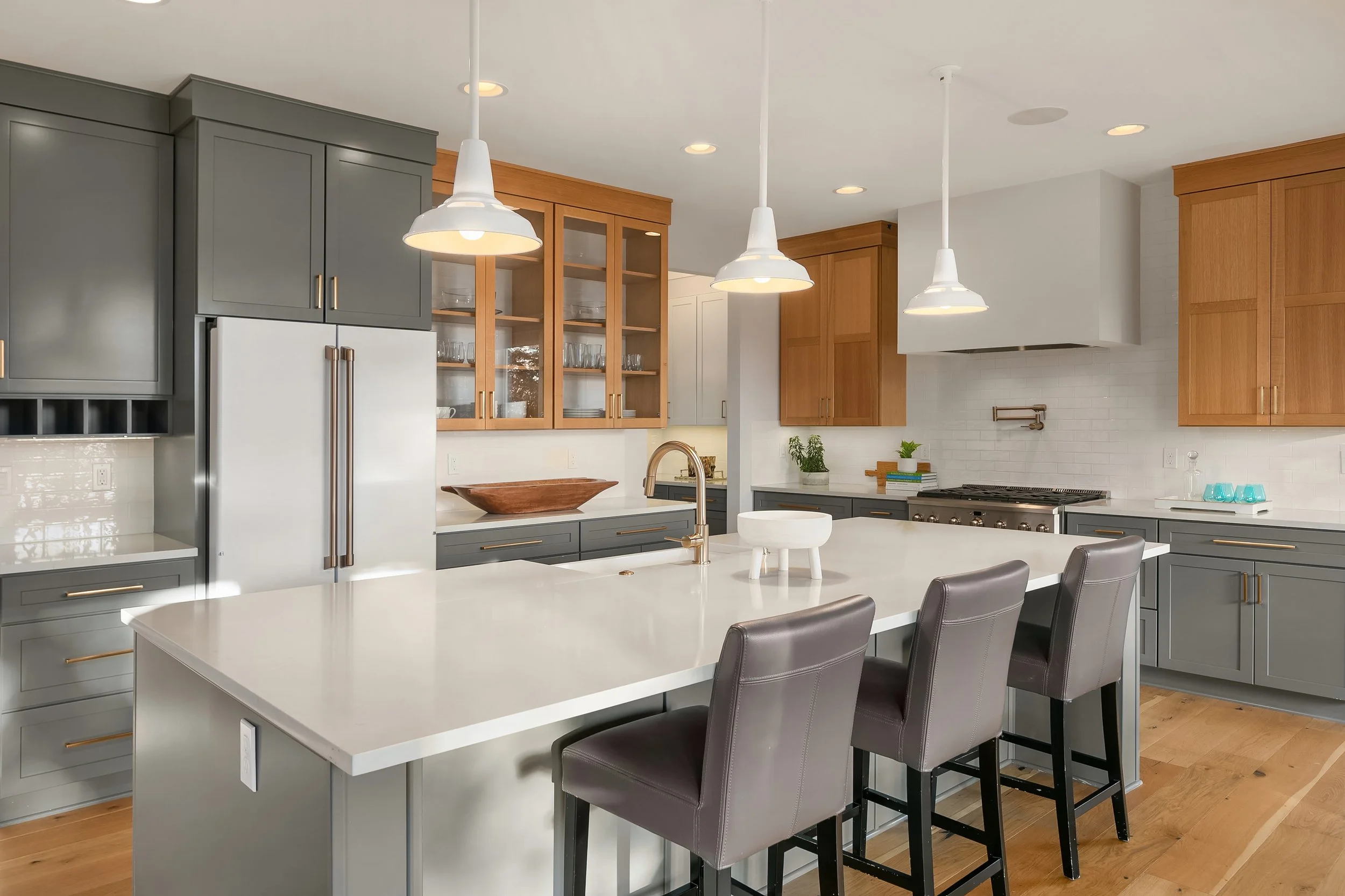 Modern kitchen with gray and wooden cabinets, white island with gray chairs, pendant lighting, stainless steel appliances, and decorative plants.