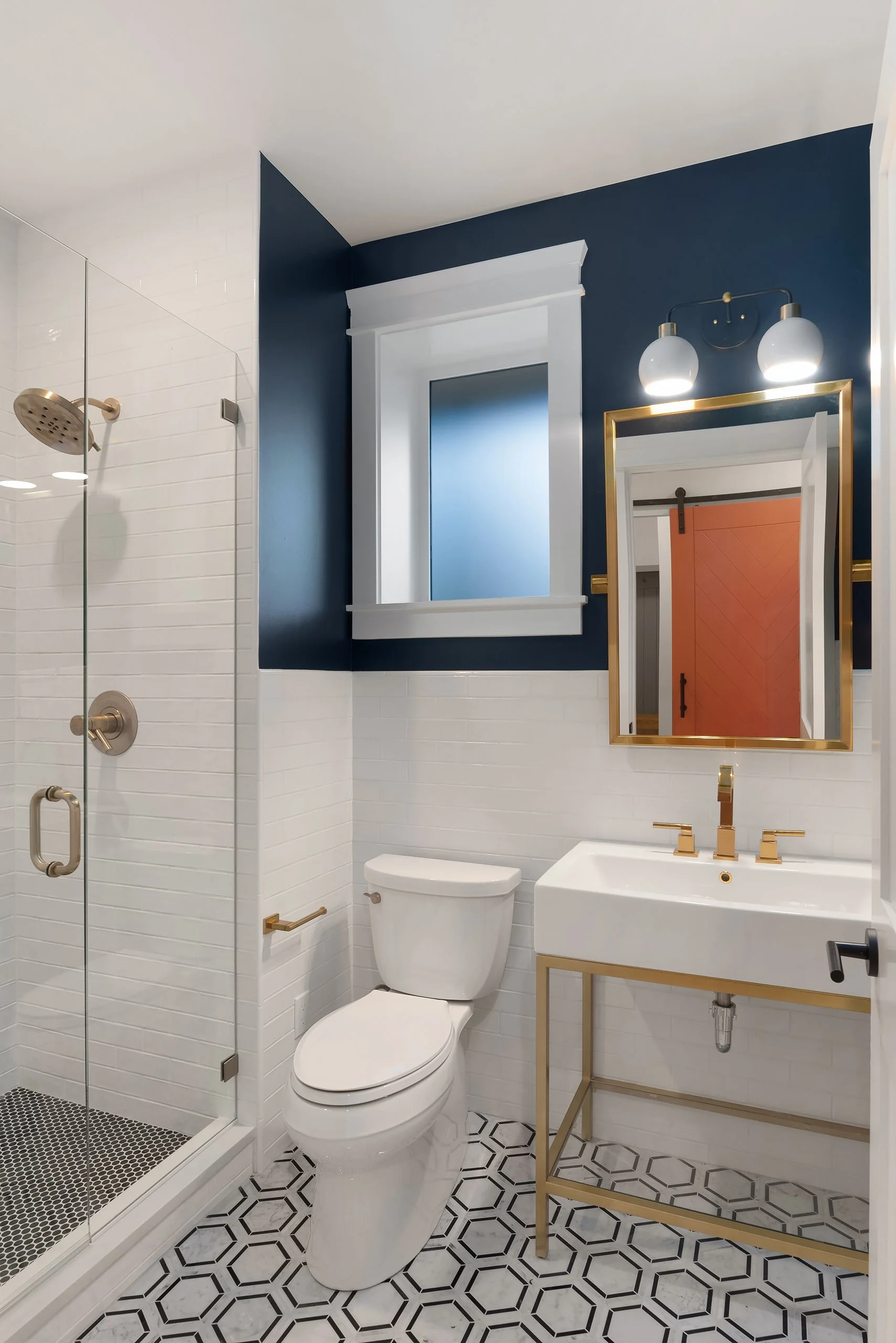 Modern bathroom with a glass-enclosed shower, white toilet, white sink with gold fixtures, and a gold-framed mirror. Blue and white walls, patterned black-and-white floor, window with white trim, and two globe-style light fixtures.