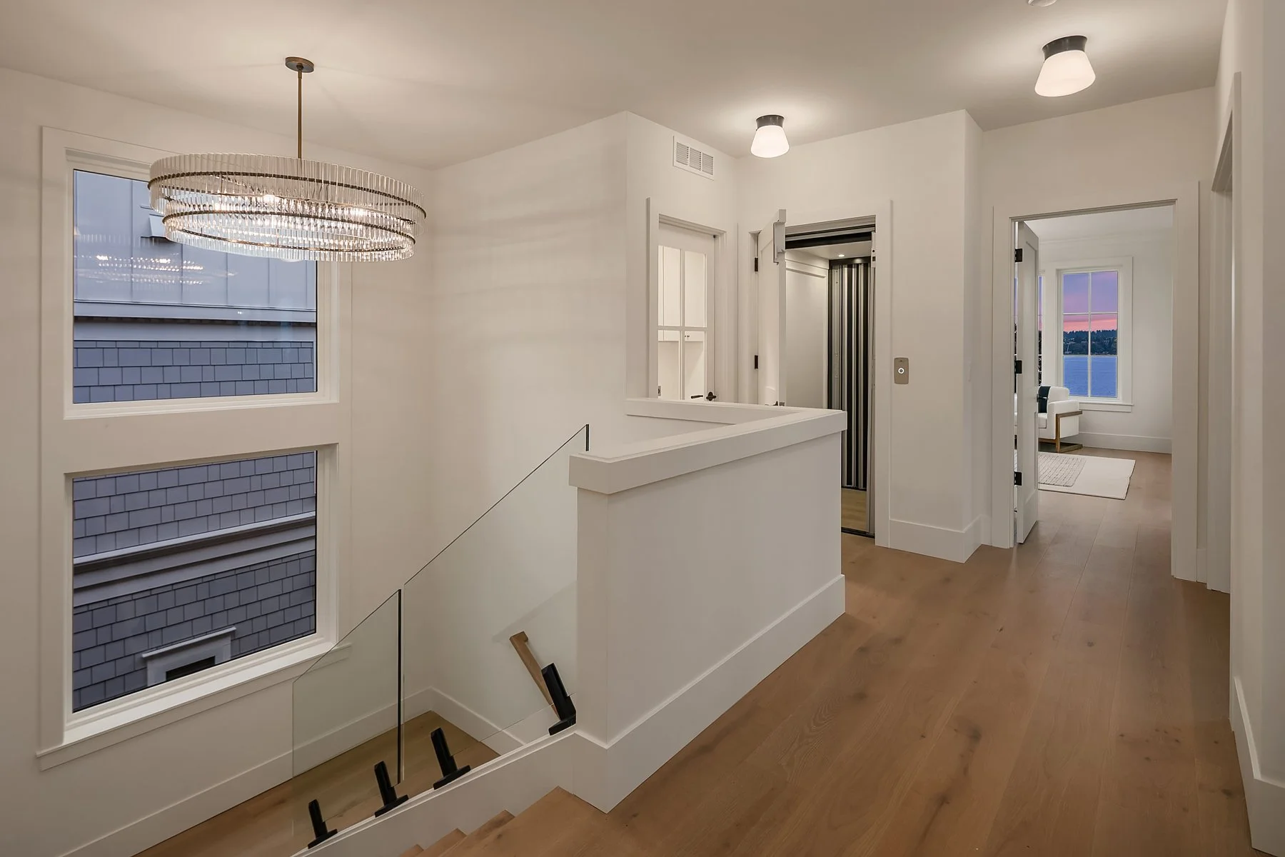 Interior view of a modern house with white walls, a staircase with glass railing, and wooden flooring. There are large windows showing a sunset sky. A chandelier hangs from the ceiling, and a hallway leads to a room with a window overlooking water.