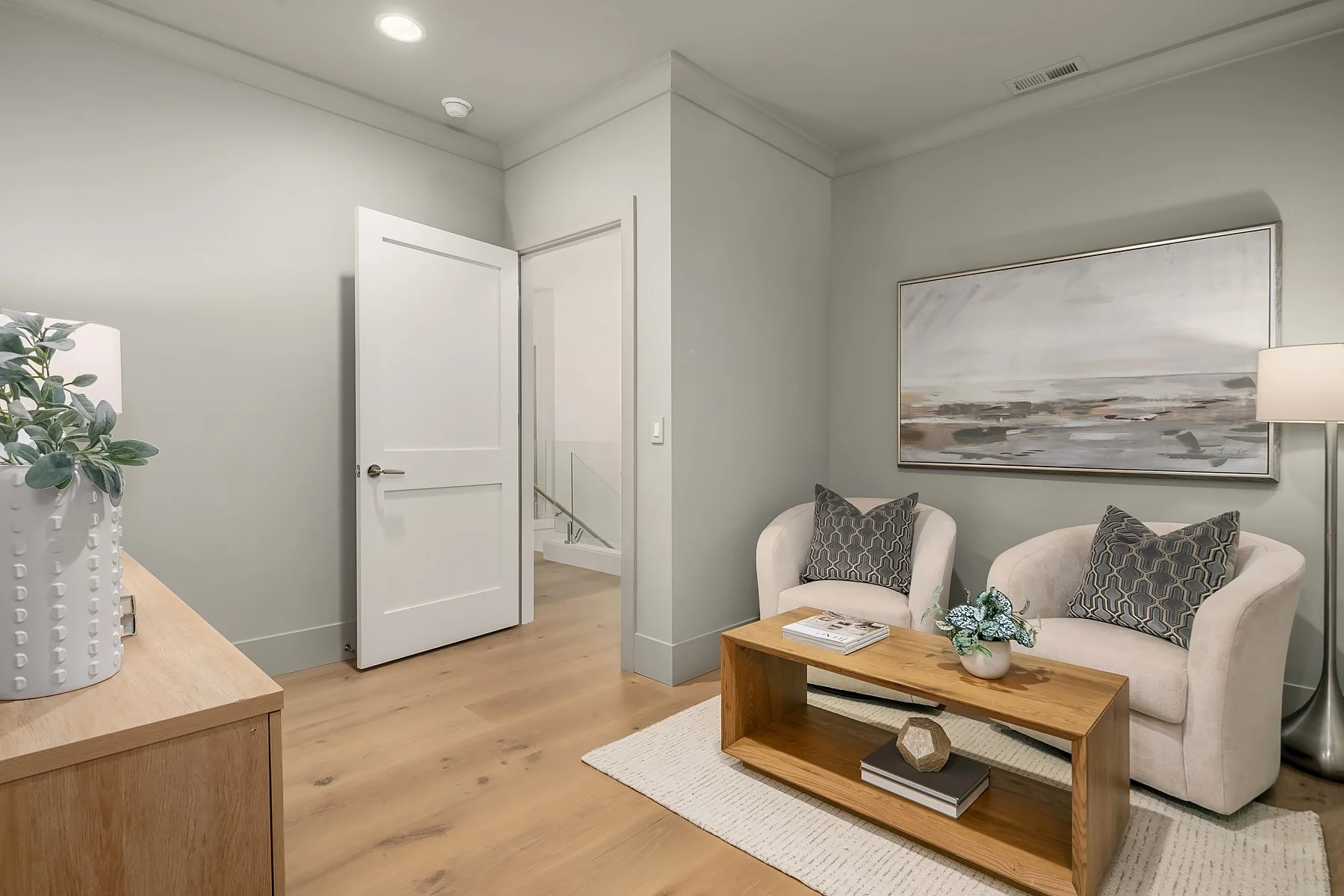 A cozy living room corner with two beige armchairs, each with a patterned pillow, a wooden coffee table with magazines and a decorative object, a large abstract painting on the wall, and a tall floor lamp. An open door leads to a staircase.