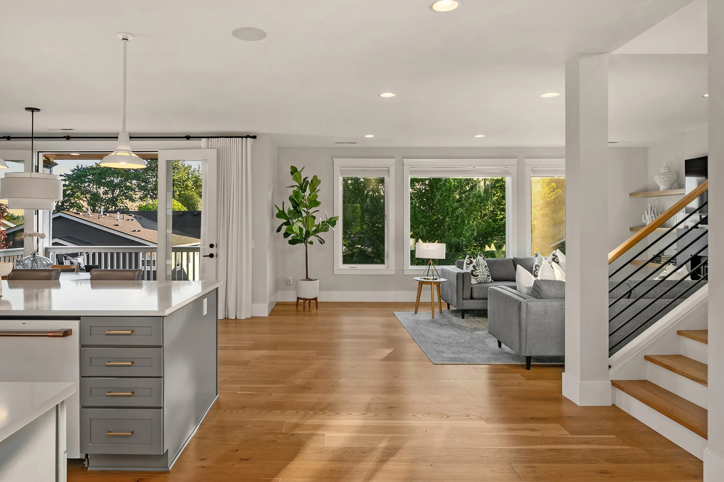 Open living room with large windows, gray sofas, wooden floors, and minimal decorations.