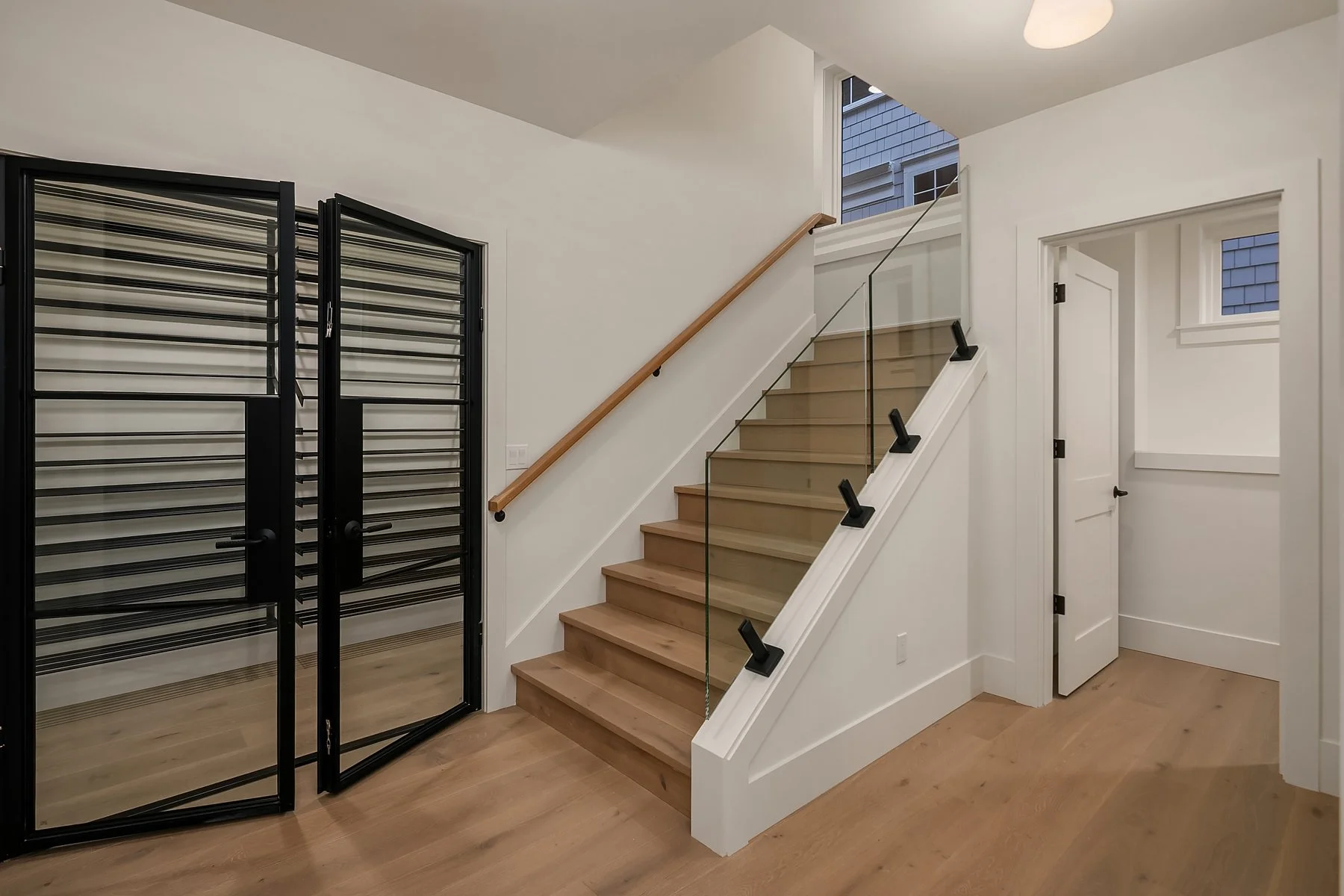 Interior of a modern home with wooden stairs, black metal and glass railing, and black double doors leading outside.