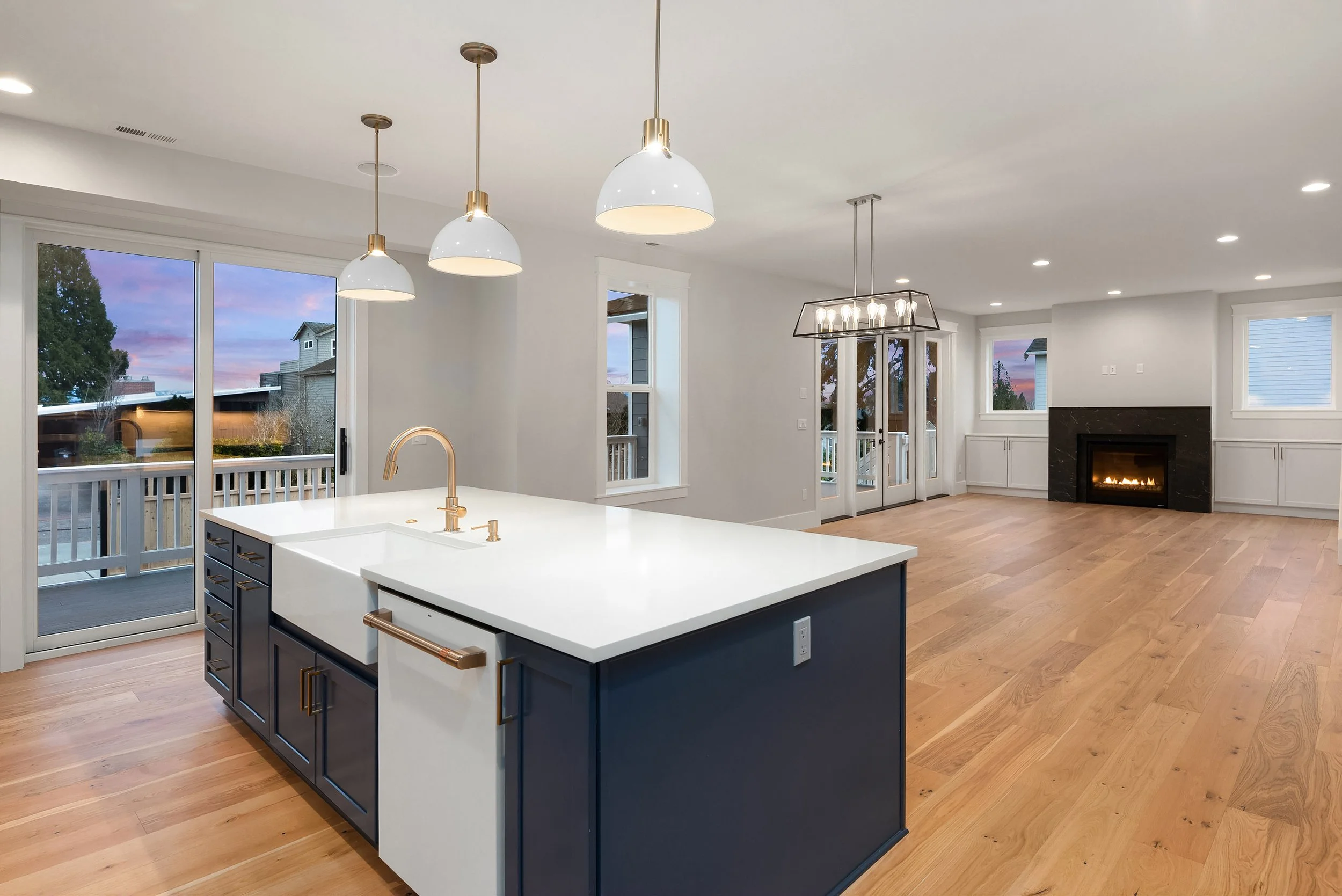 Modern open-concept living room and kitchen with wood flooring, white walls, large windows, a blue kitchen island with brass hardware, pendant lights, a chandelier over the dining area, and a fireplace