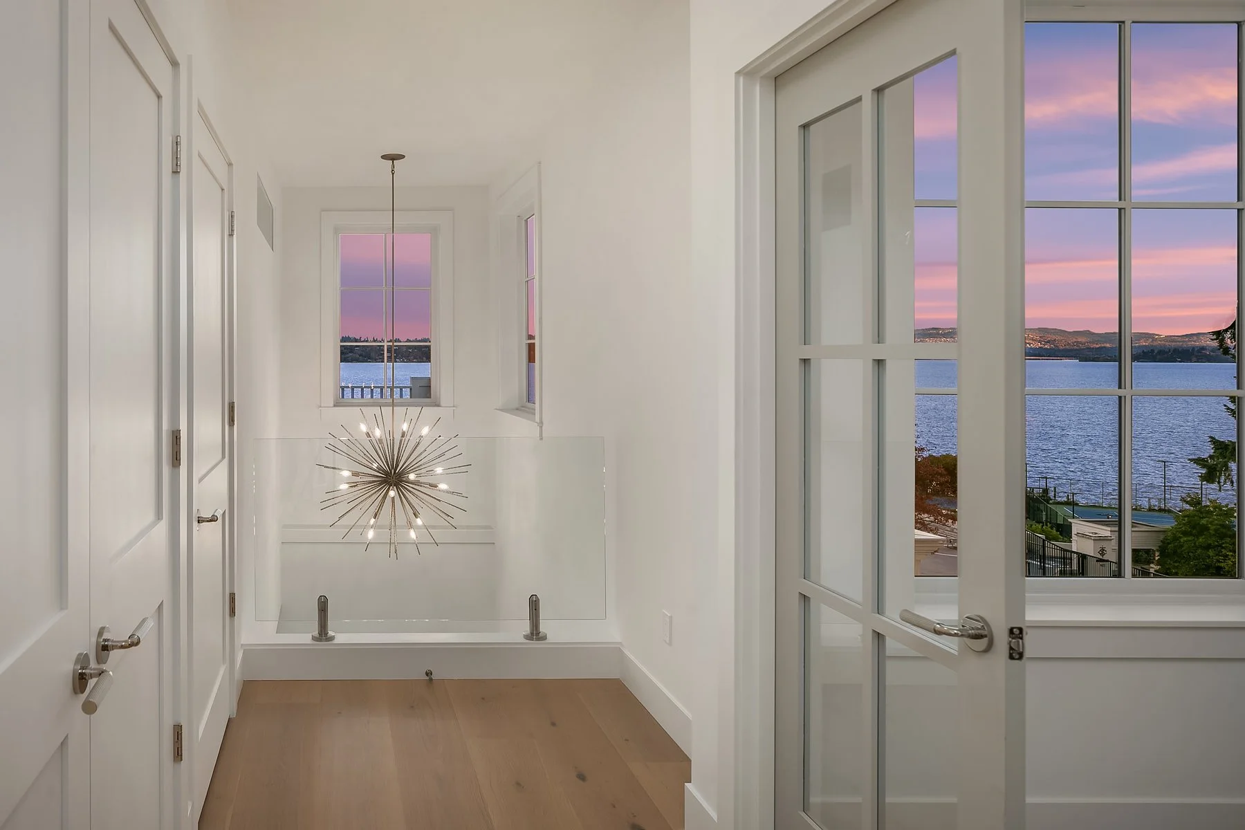 Interior view of a bright, white room with large windows looking out over a body of water and a scenic sunset sky.