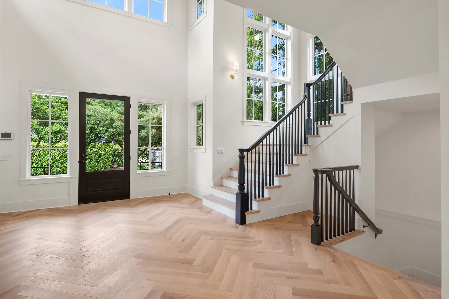 Bright entrance hall with large windows, black door, and staircase with black railing and wooden steps.