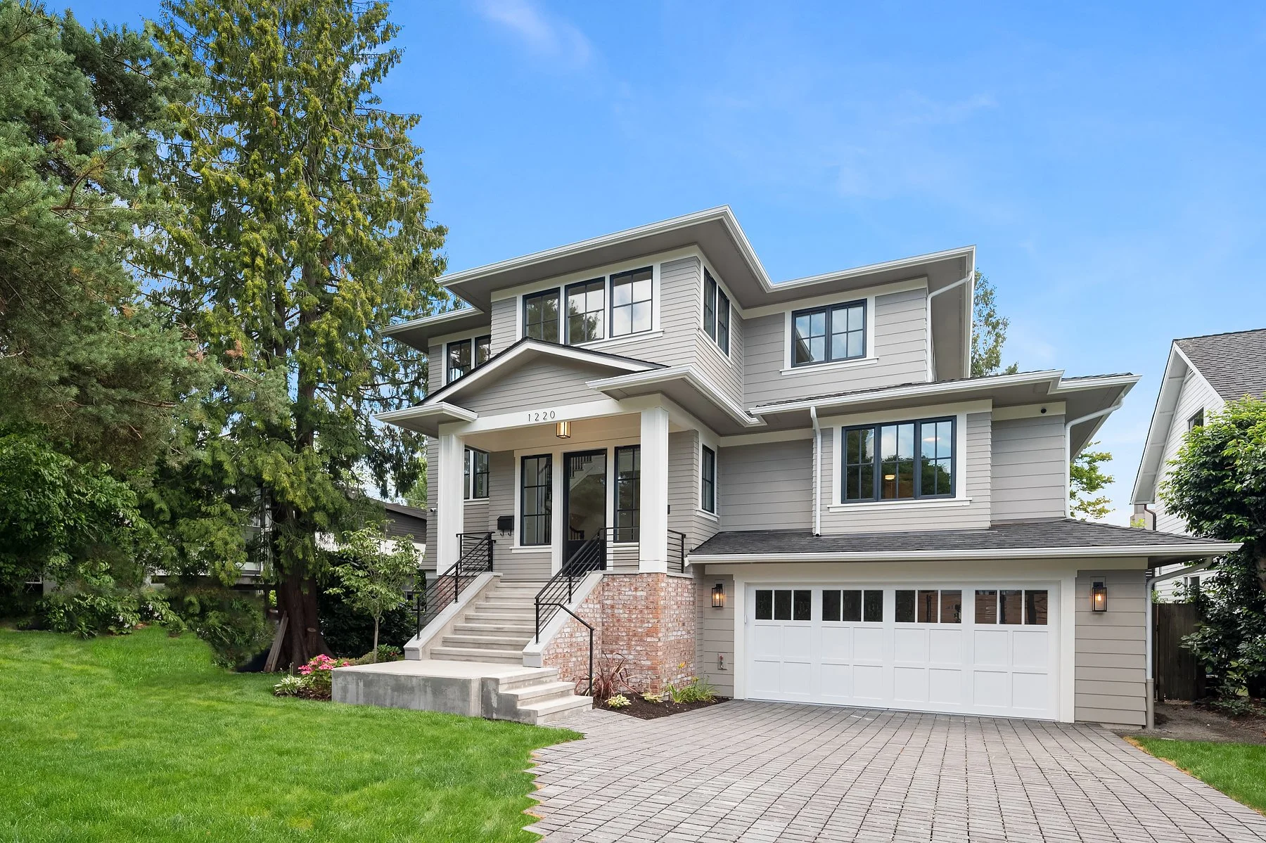 A modern, multi-story house with gray siding, large windows, and a white garage door, surrounded by a green lawn and trees under a clear blue sky.