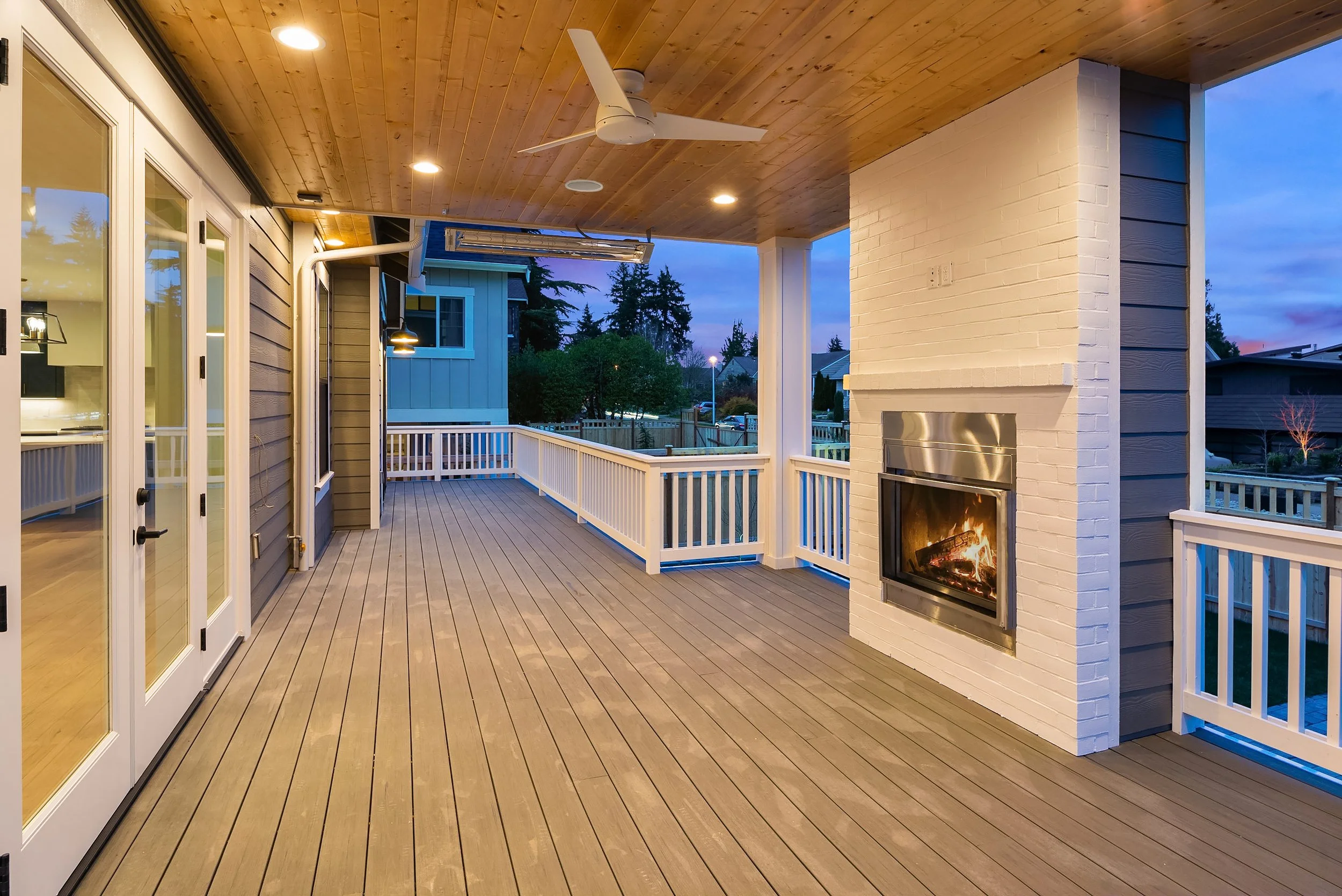 A spacious outdoor porch with wooden flooring, white railing, a white brick fireplace with a fire inside, a ceiling fan, and recessed lighting. The porch is adjacent to a house with sliding glass doors, and in the background, there are trees and neig