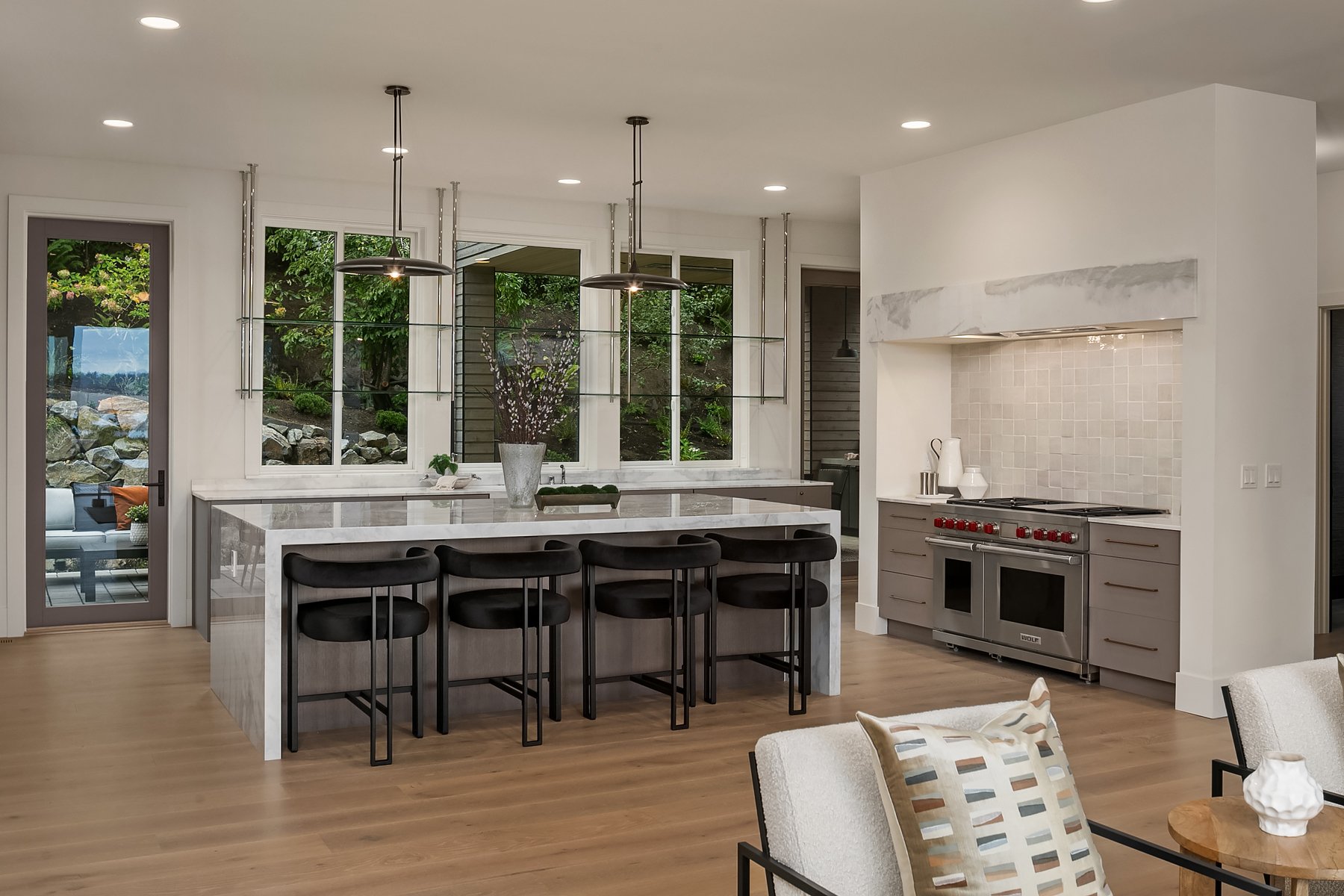 Modern kitchen with white walls, large windows, marble island, black chairs, and a stainless steel range.
