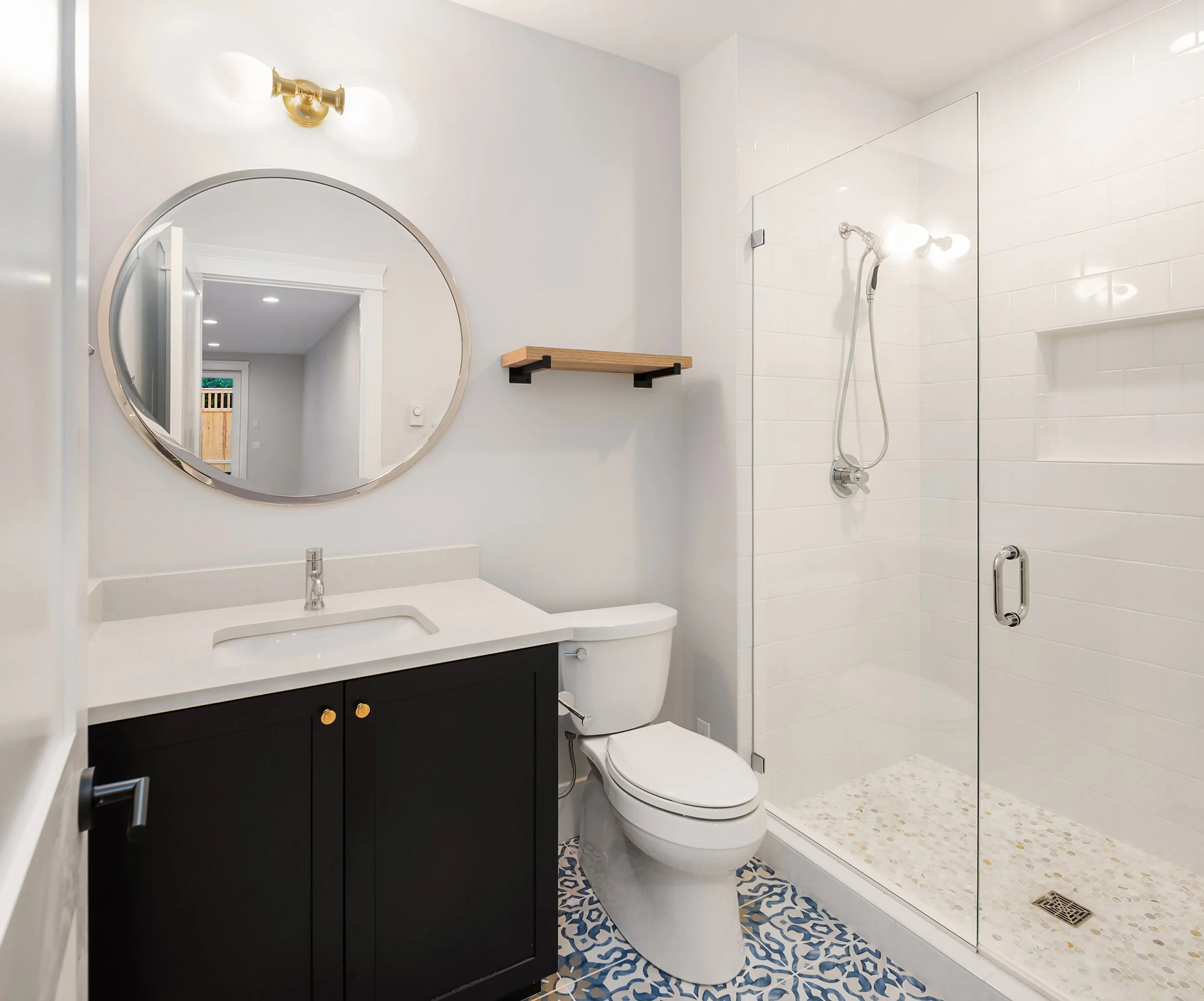 Modern bathroom with a black vanity, white countertop, round mirror, white toilet, and a glass-enclosed walk-in shower with white tile walls and pebble floor.