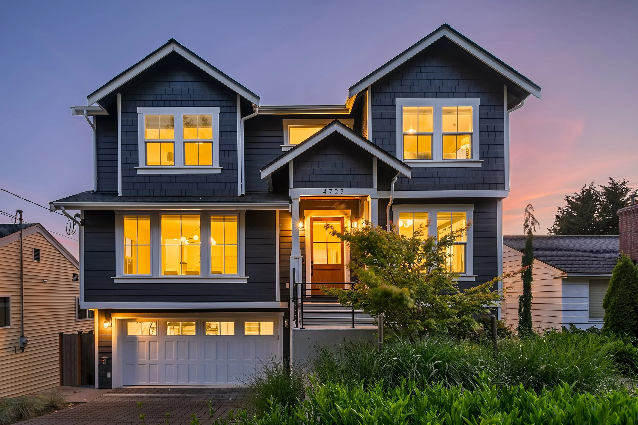 A two-story house with blue siding and multiple large windows, illuminated from within, during sunset. The house has a garage at the front, with a small staircase leading to the main entrance, and surrounded by greenery.