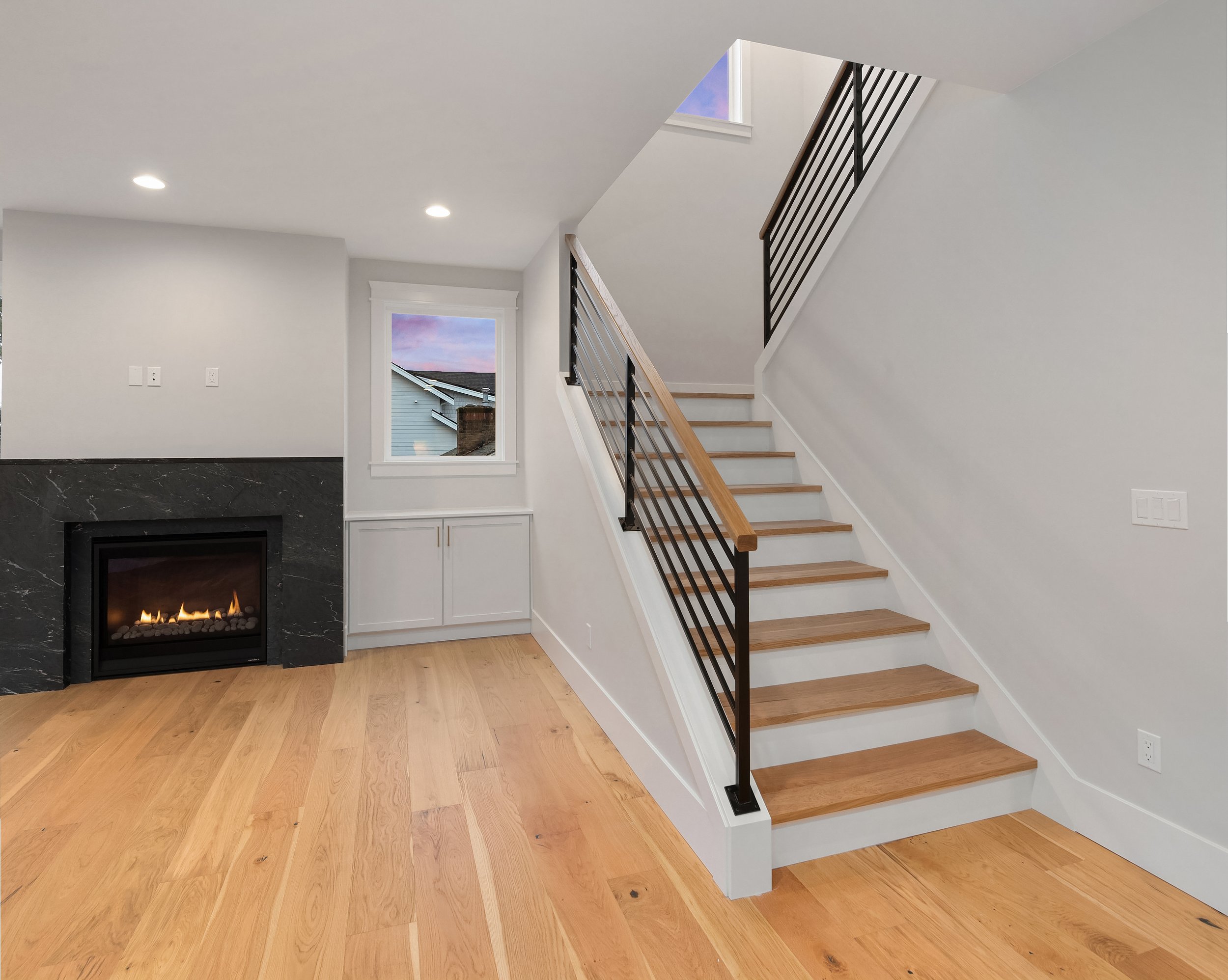 Interior of a modern living space with a staircase, fireplace, white walls, hardwood flooring, a window, and recessed lighting.