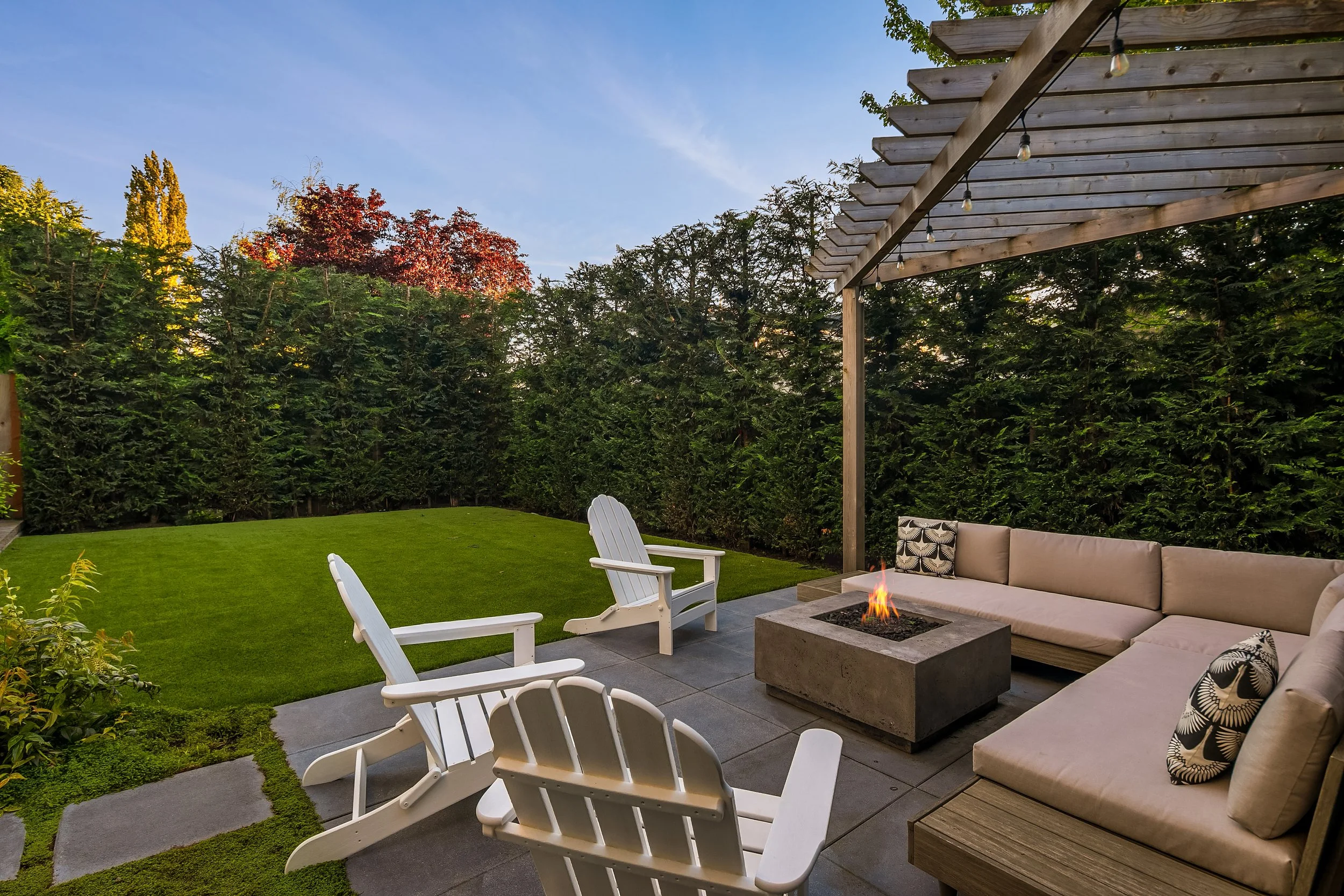 Backyard patio with fire pit, beige sectional sofa with pillows, white Adirondack chairs, lush green lawn, tall hedges, and a pergola with string lights in the evening.