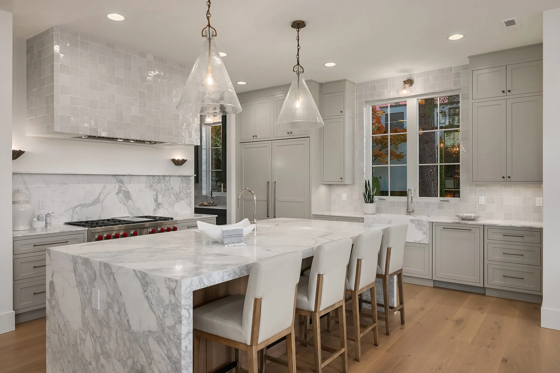 Modern kitchen with white marble island, gray cabinets, tile backsplash, pendant lights, and large windows
