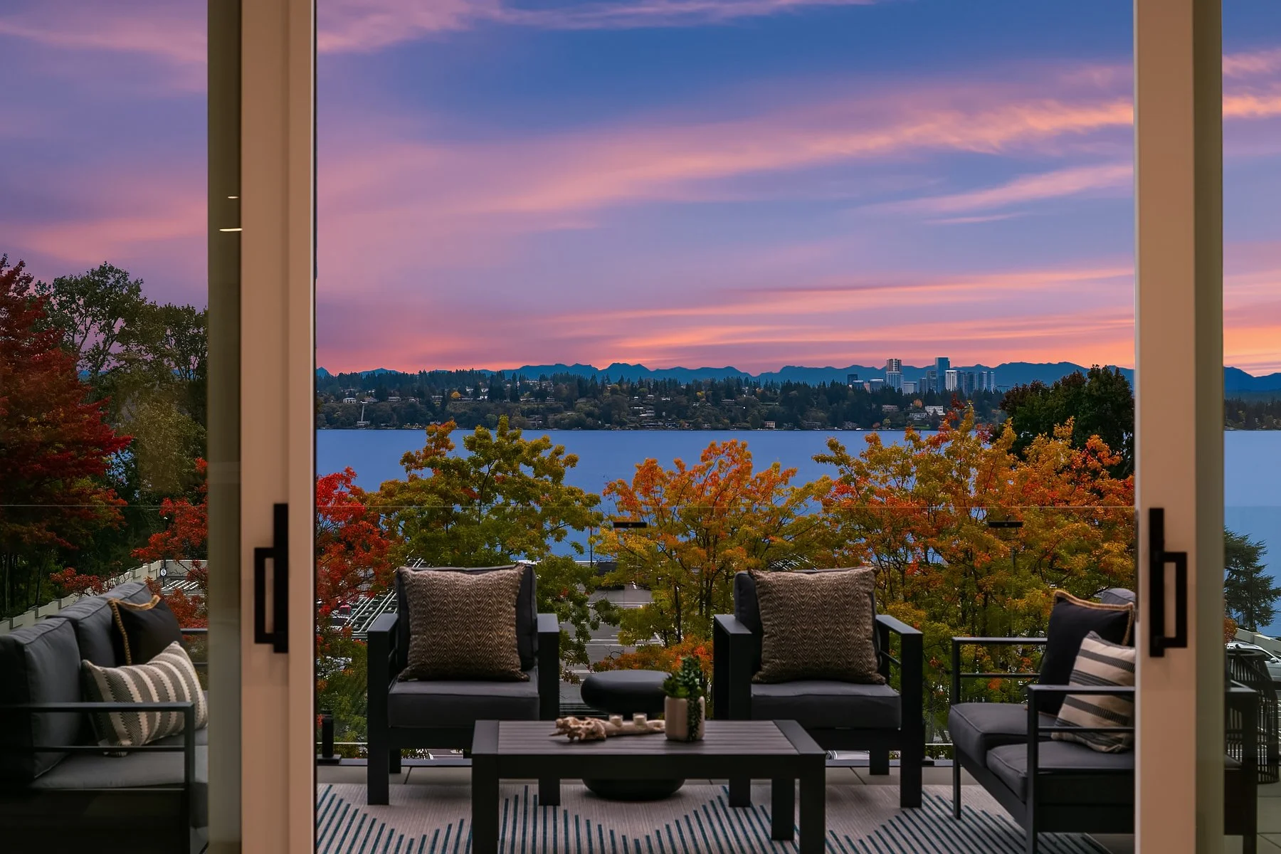 A view of a lake and colorful autumn trees from a balcony with black outdoor furniture and decorative pillows at sunset