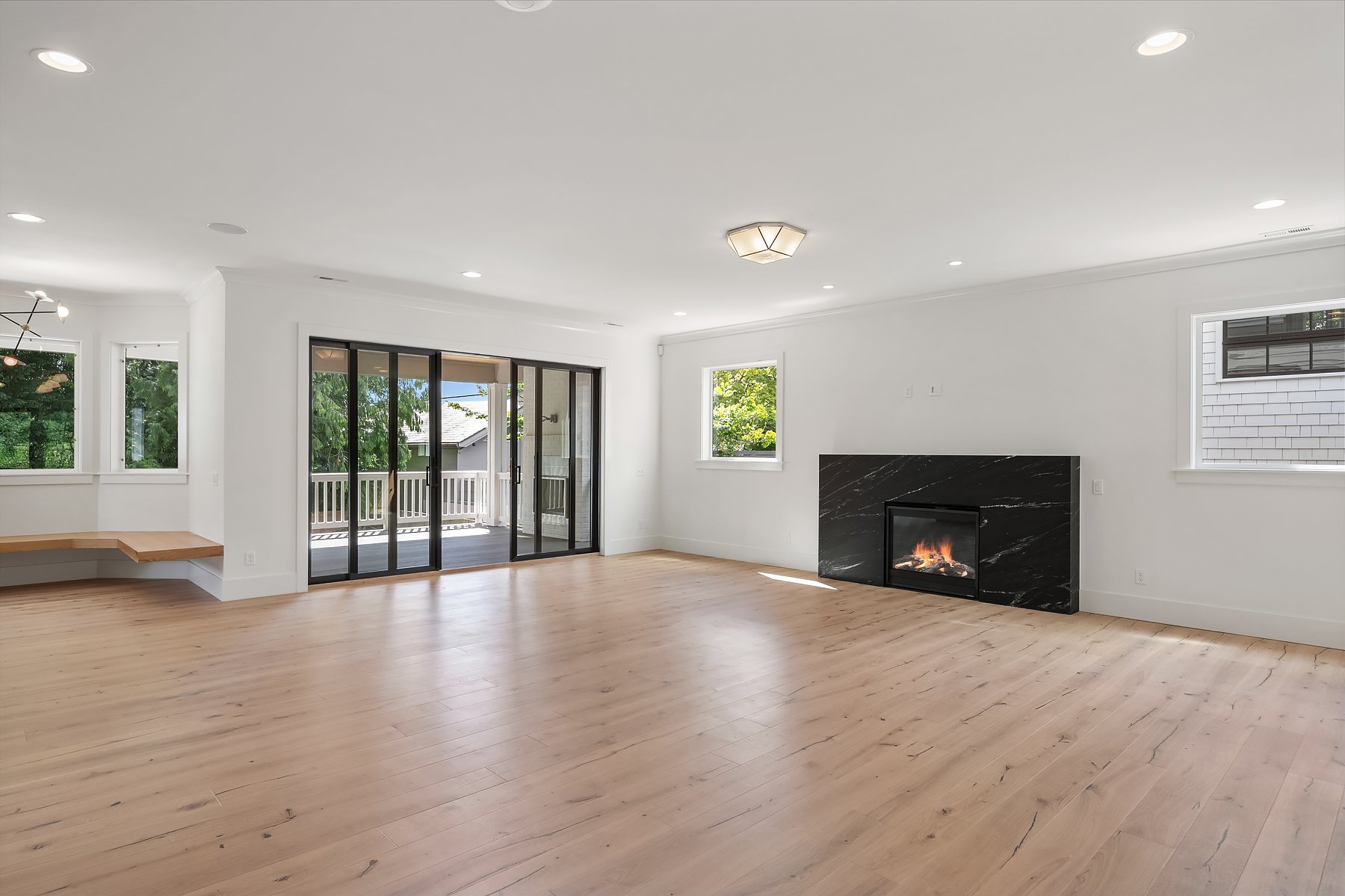 Empty living room with light wood flooring, white walls, a black marble fireplace, multiple windows, and a sliding glass door leading to a balcony.
